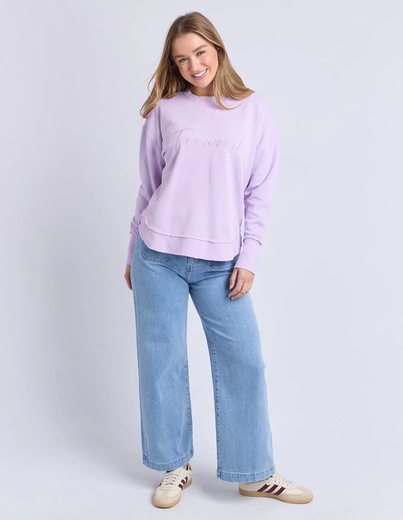 Image of Woman wearing a Foxwood light purple sweatshirt and blue jeans on a white background