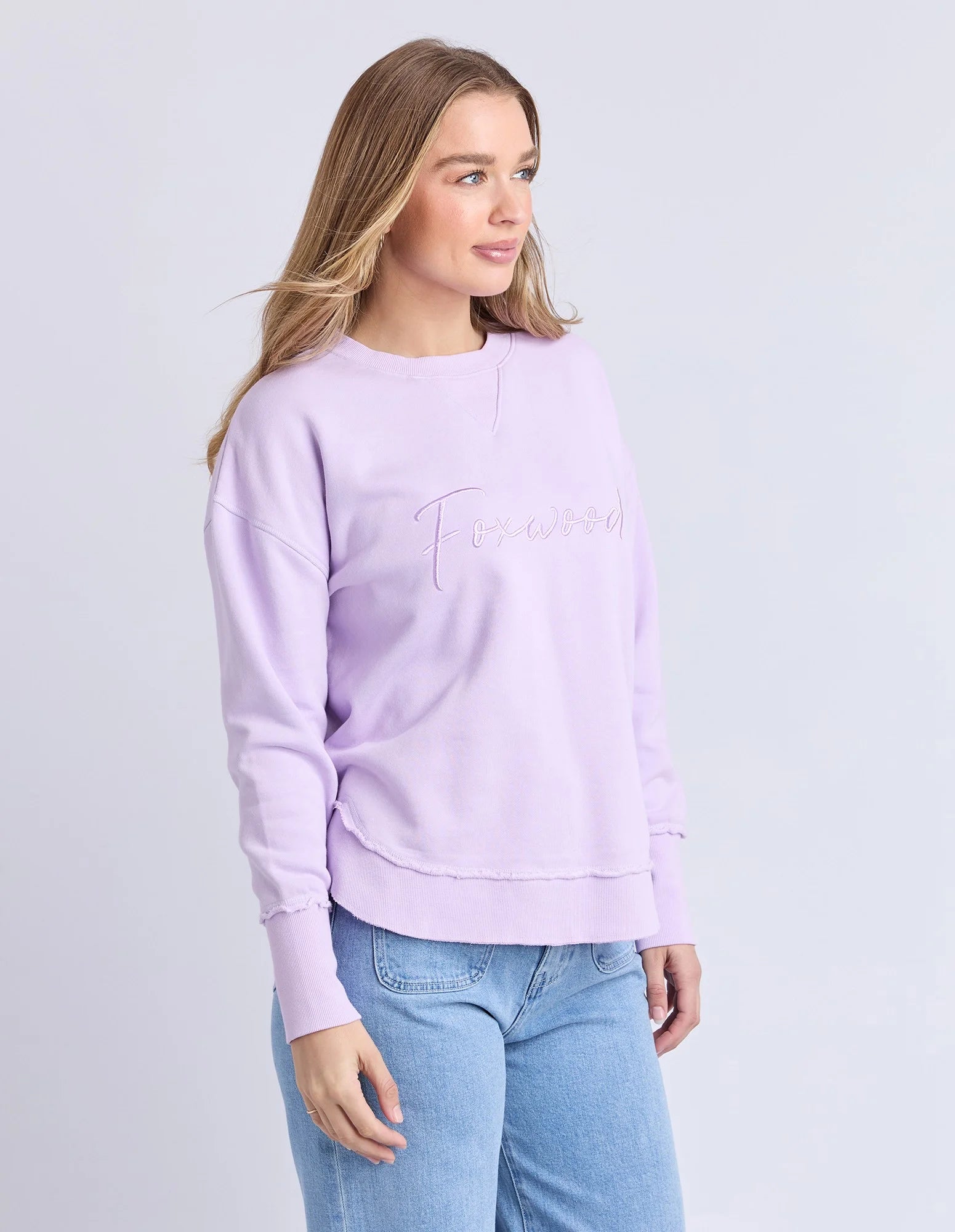 Woman wearing a Foxwood light purple sweatshirt and blue jeans on a plain background