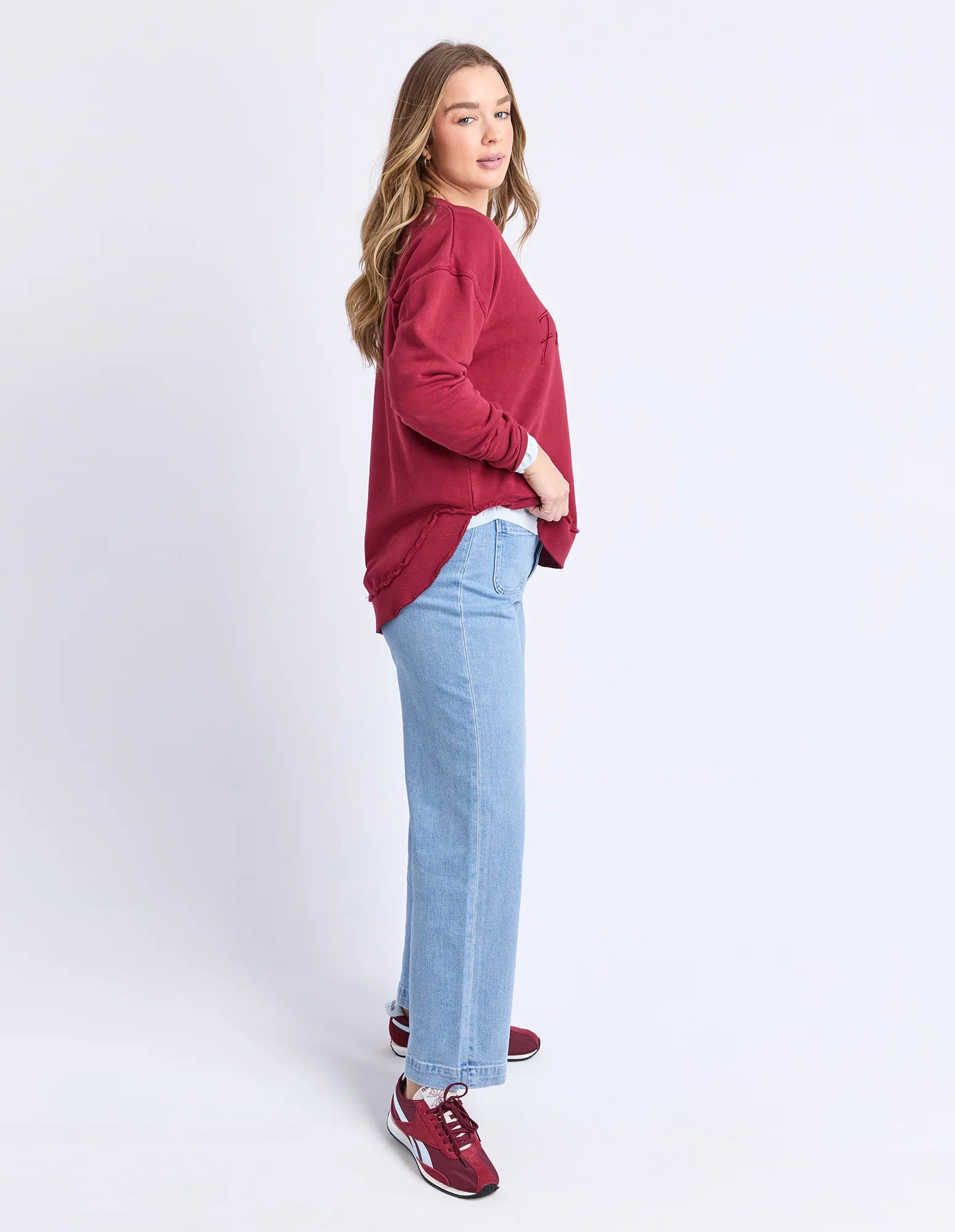 Woman wearing Foxwood signature crew in rhubarb paired with denim jeans on a plain background