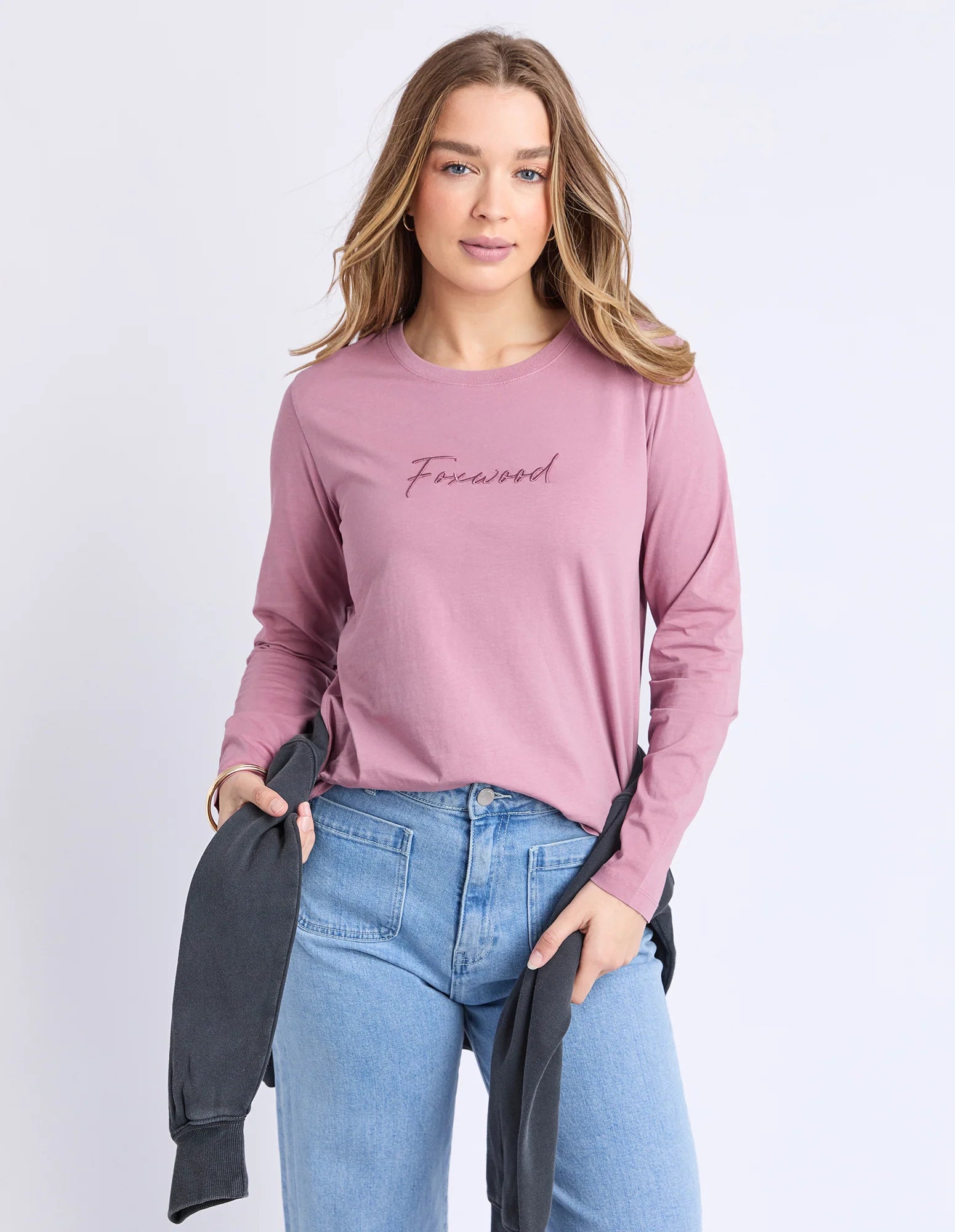 Image of Woman wearing a pink sweatshirt with 'Foxwood' text, holding a dark jacket against a light purple background
