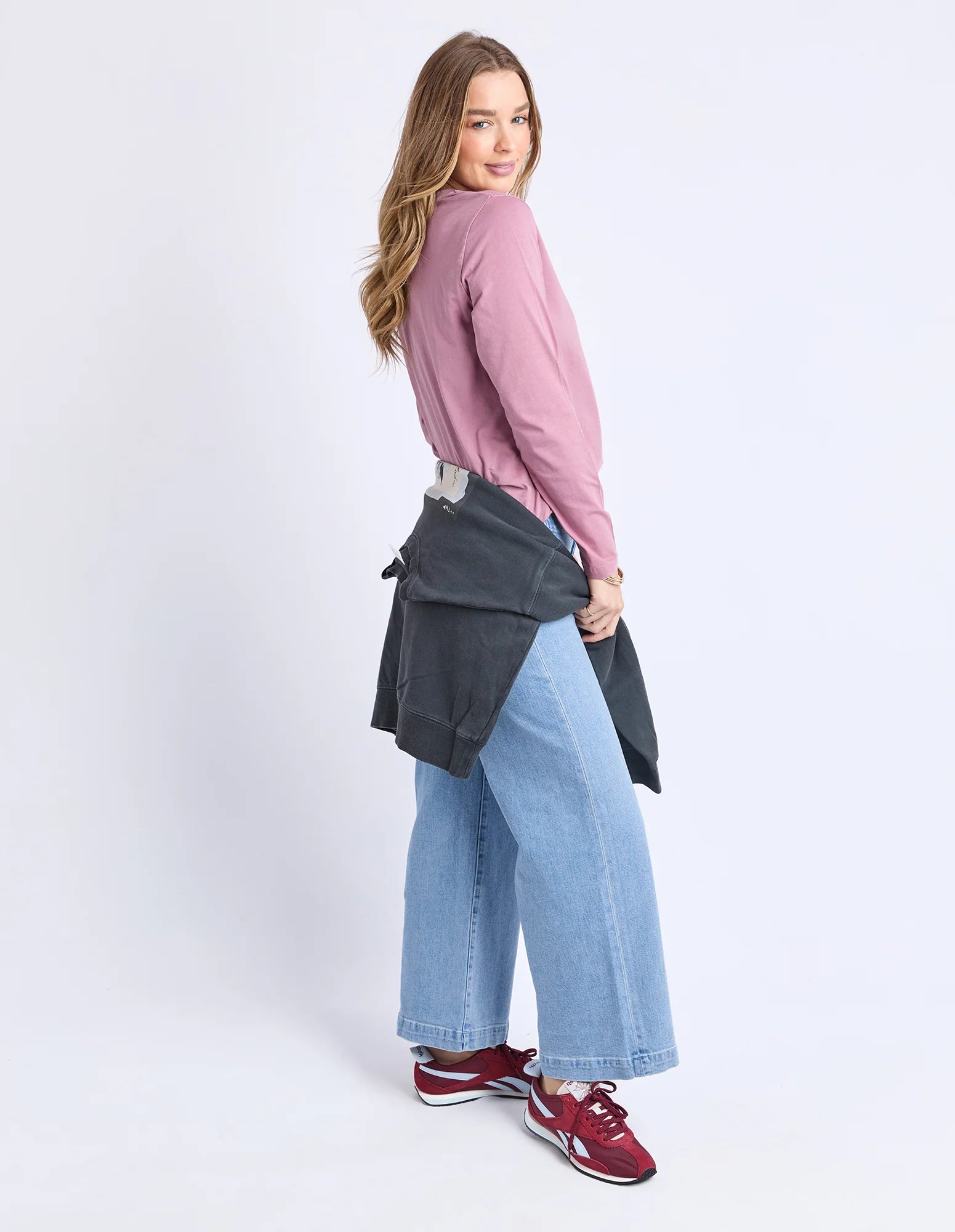 Woman wearing a Foxwood pink shirt, blue jeans, and red sneakers holding a gray bag on a white background
