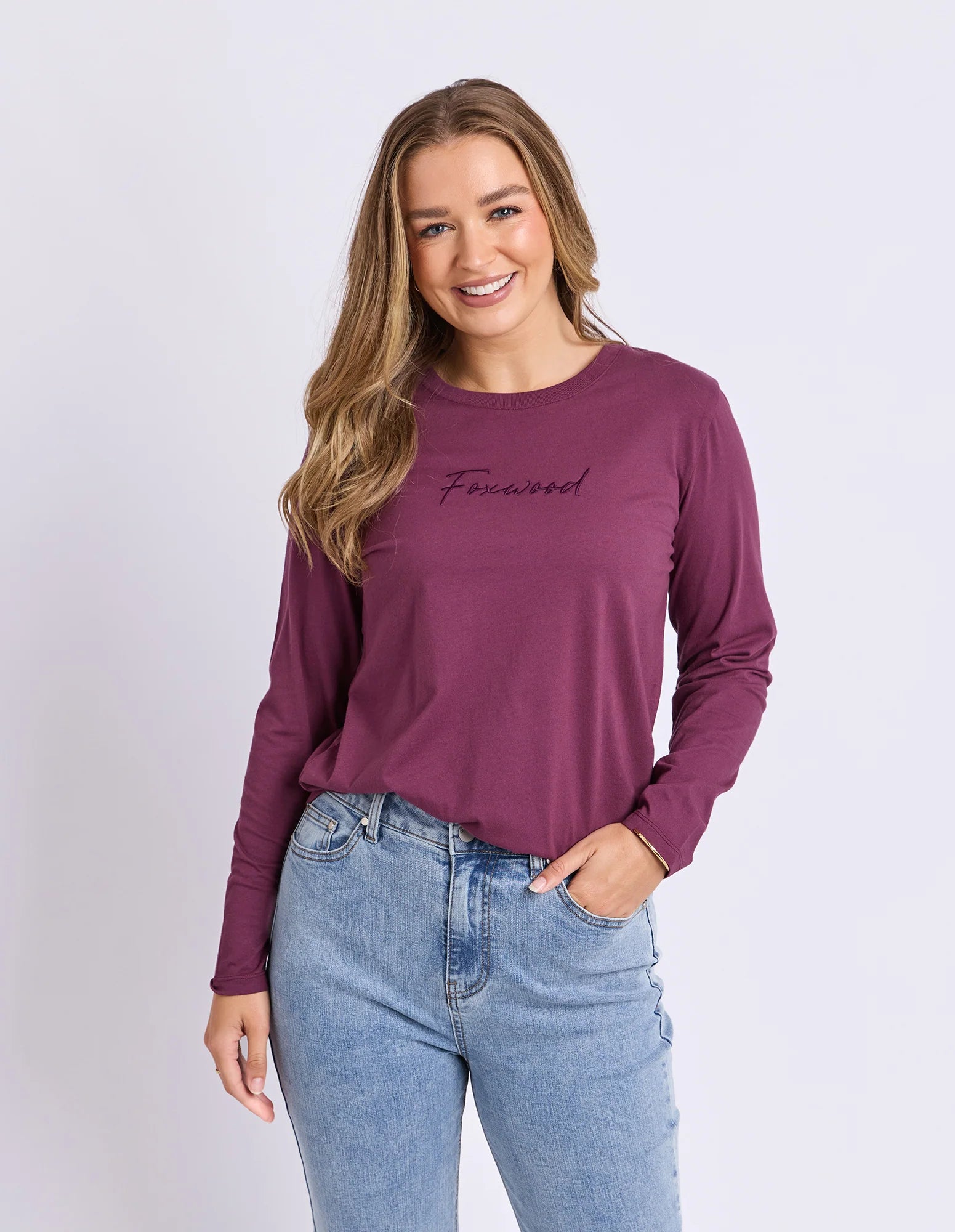 Image of A woman modeling a Foxwood long-sleeve crewneck top in a deep plum purple color, featuring the word 'Finewood' embroidered in black script across the chest. She is wearing light-wash blue jeans against a clean, light gray studio background.