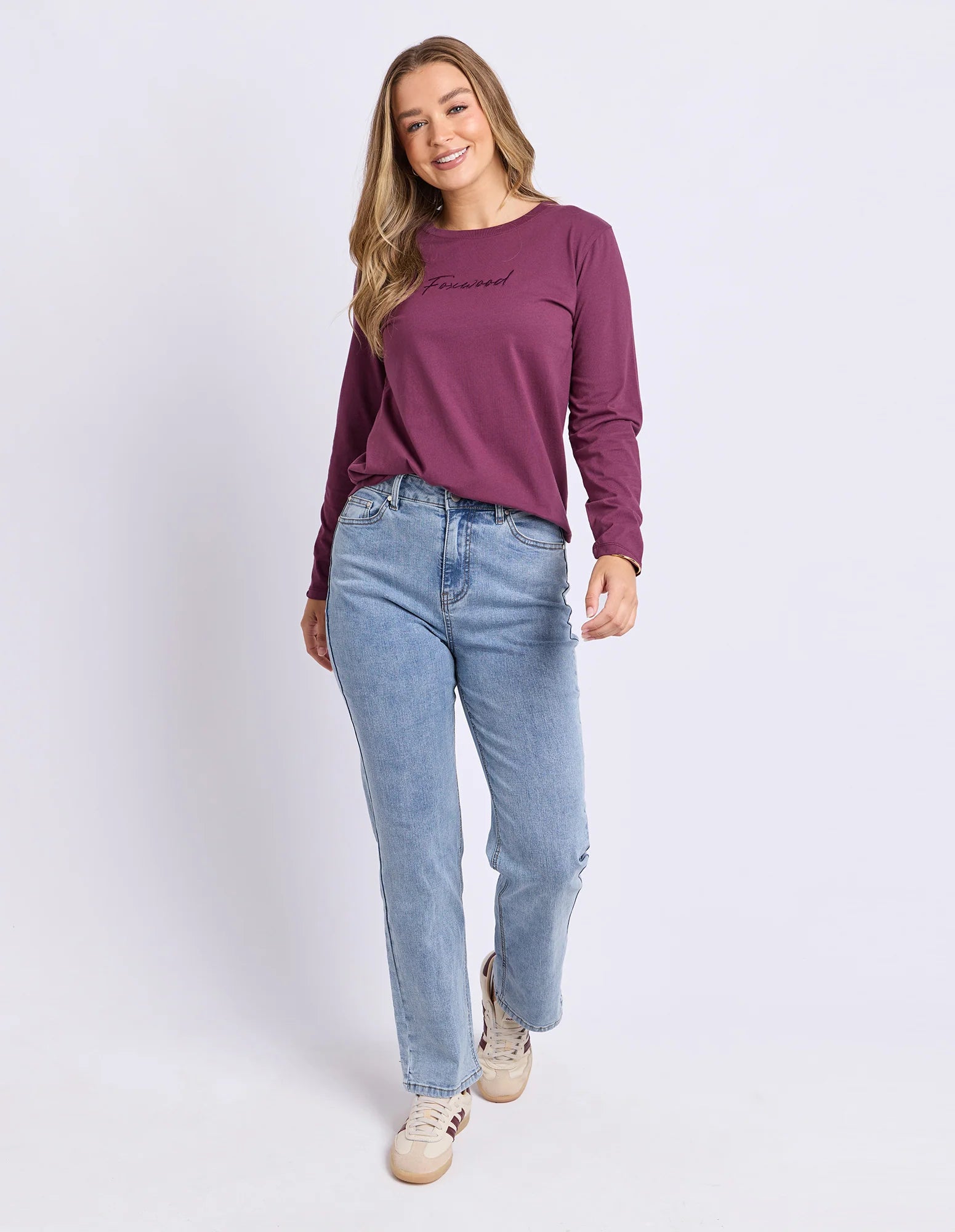 Image of A woman modeling a Foxwood long-sleeve crewneck top in deep plum purple, paired with light-wash blue jeans and beige sneakers, against a plain light gray studio background.