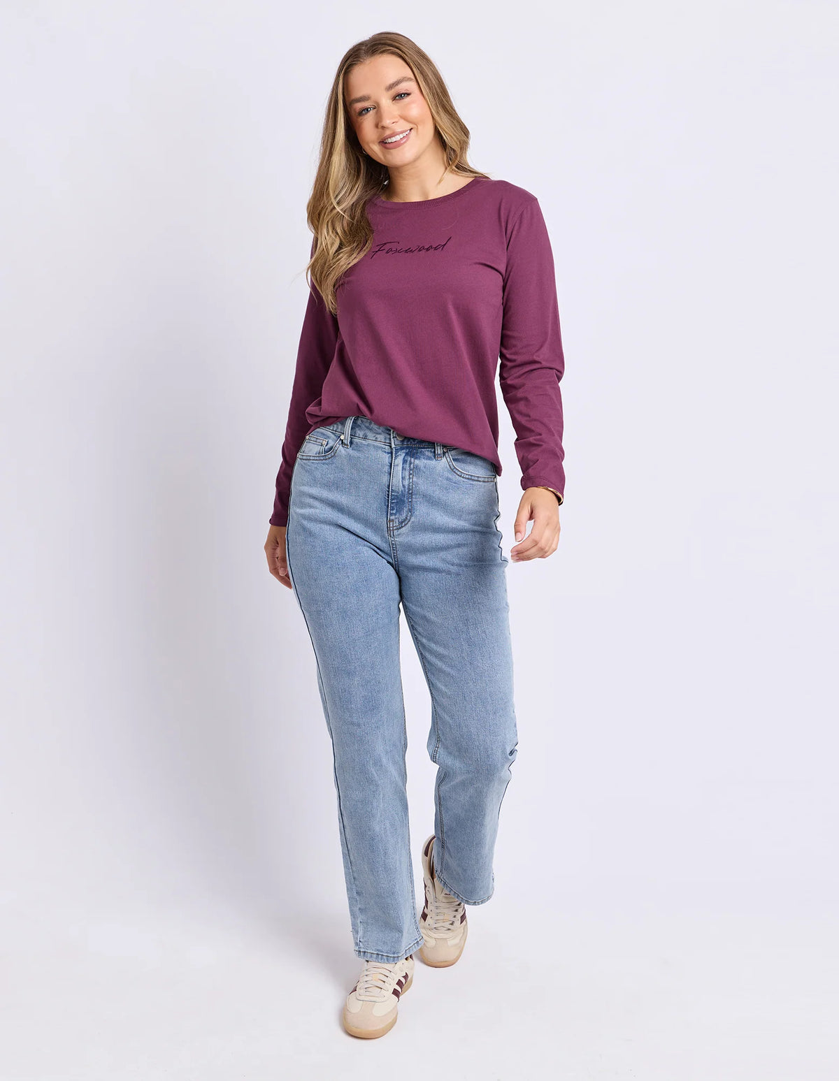 A woman modeling a Foxwood long-sleeve crewneck top in deep plum purple, paired with light-wash blue jeans and beige sneakers, against a plain light gray studio background.