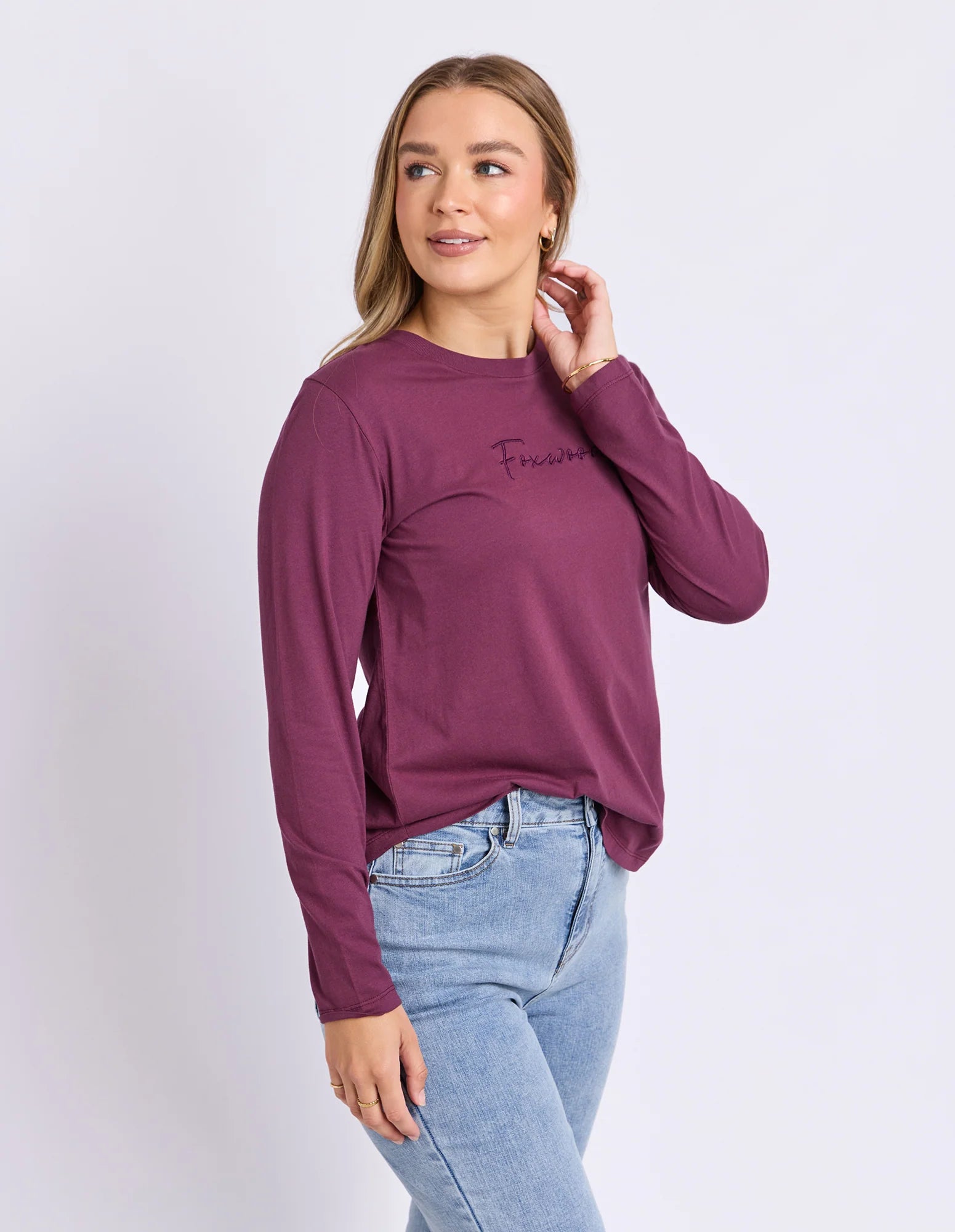 Image of A woman modeling a Foxwood long-sleeve crewneck top in a deep plum purple color, featuring a small black script logo on the left chest, paired with light blue jeans against a plain light gray background.