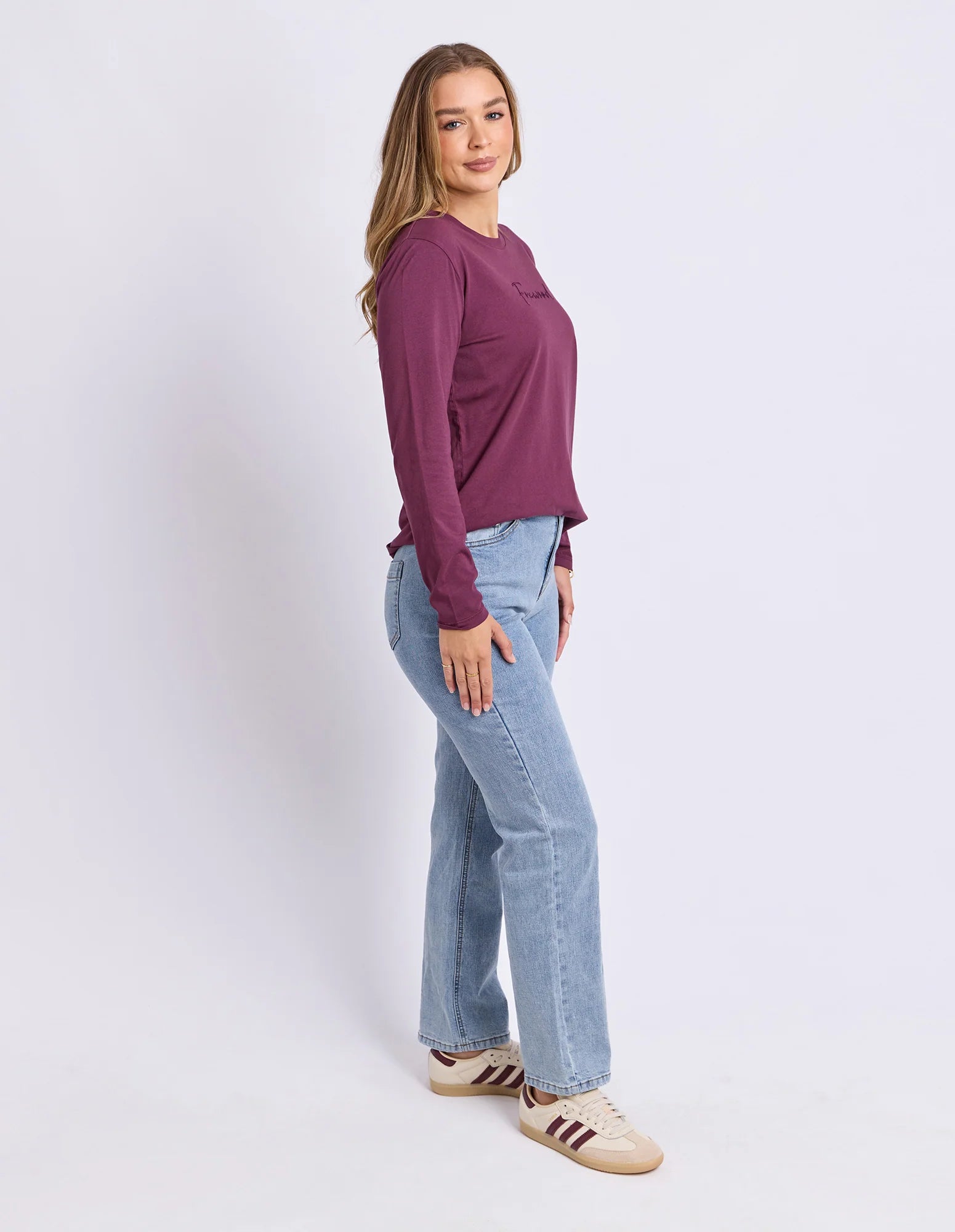 Image of A woman modeling a Foxwood long-sleeve crewneck top in deep plum purple, paired with light-wash blue jeans and beige sneakers, standing against a plain light gray studio background.