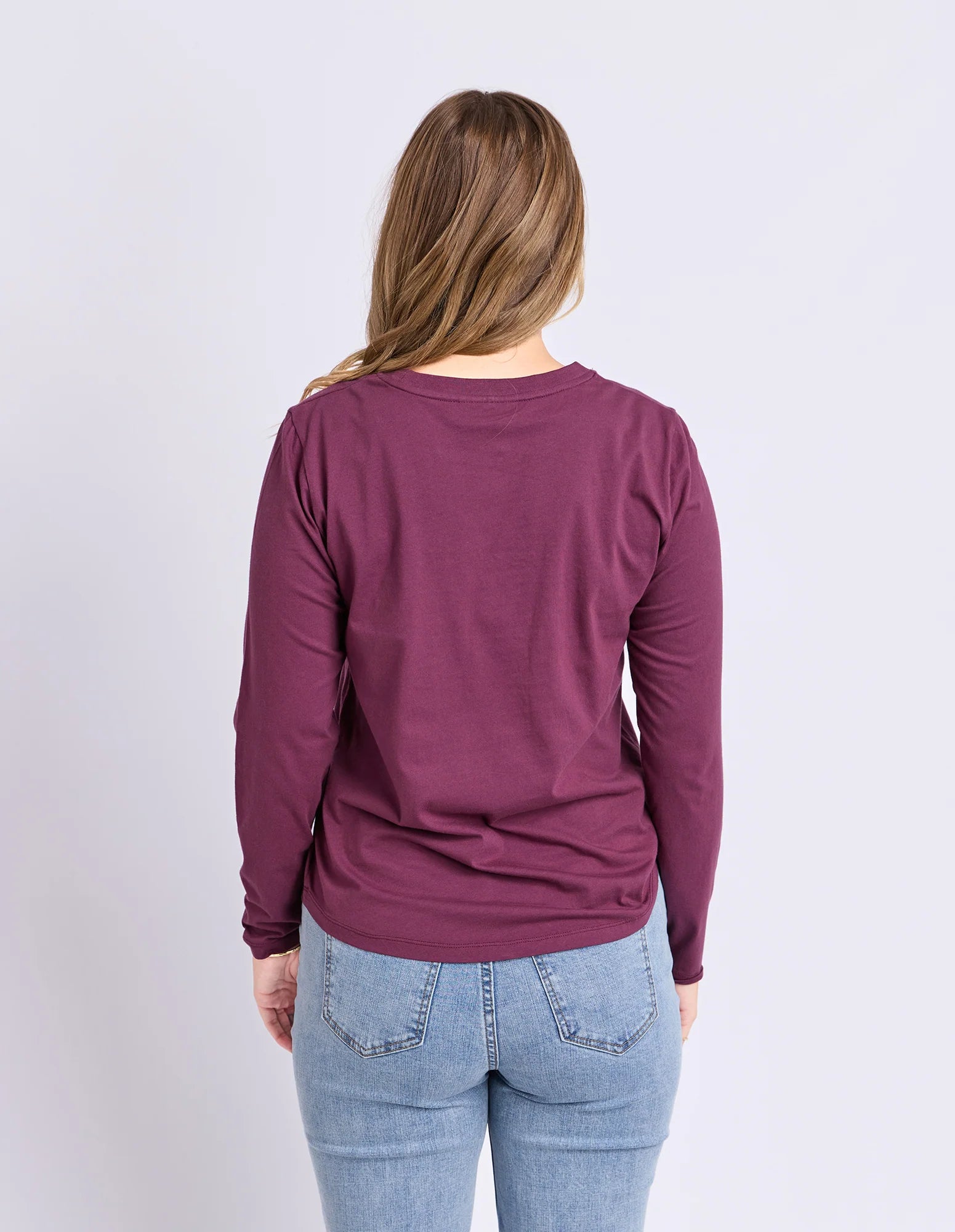 Image of A woman viewed from the back wearing a Foxwood long-sleeve crewneck top in a deep plum purple color, paired with light blue jeans, against a plain light gray background.
