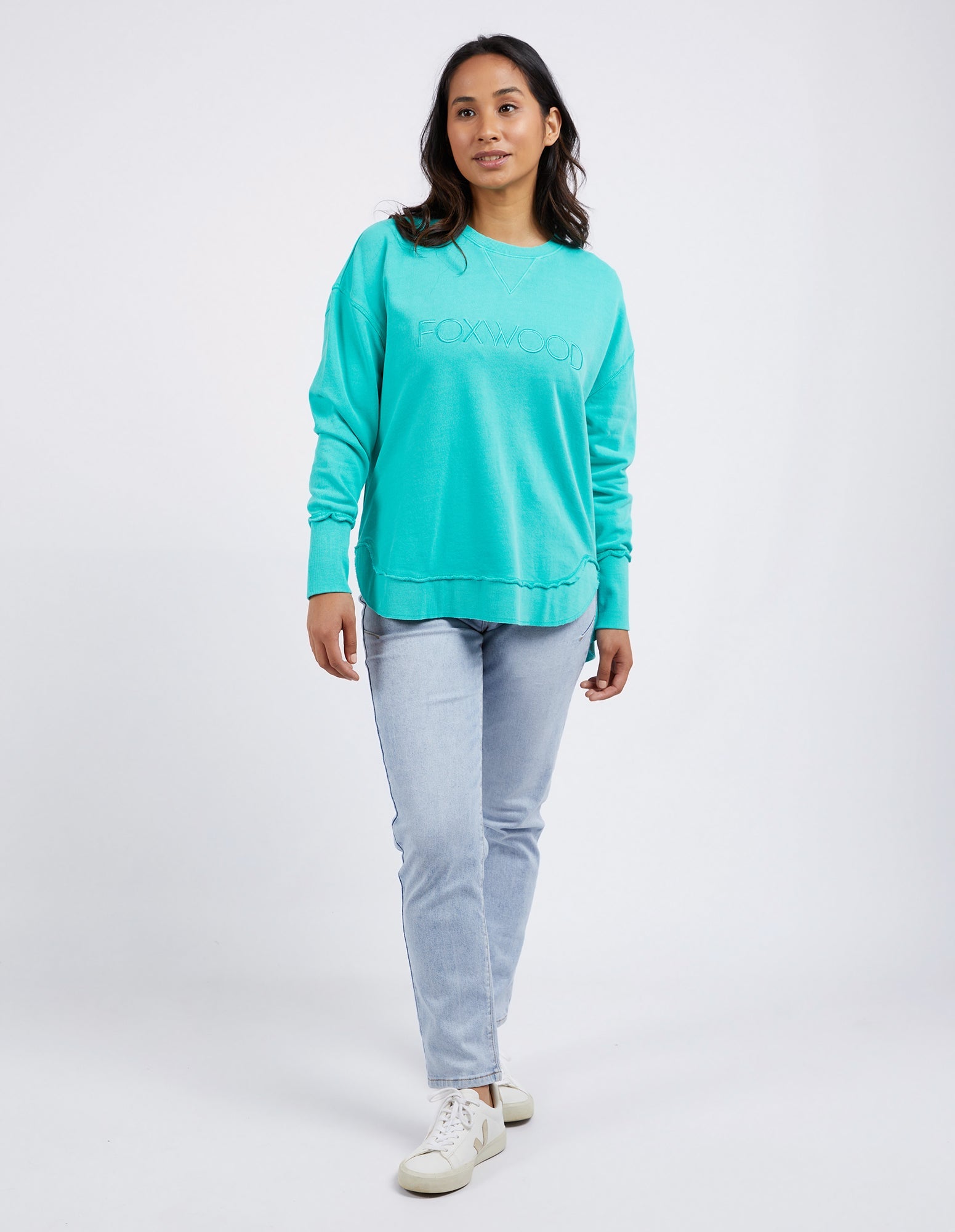 Woman wearing Foxwood Simplified crew in a teal colourway paired with denim jeans on a plain background 