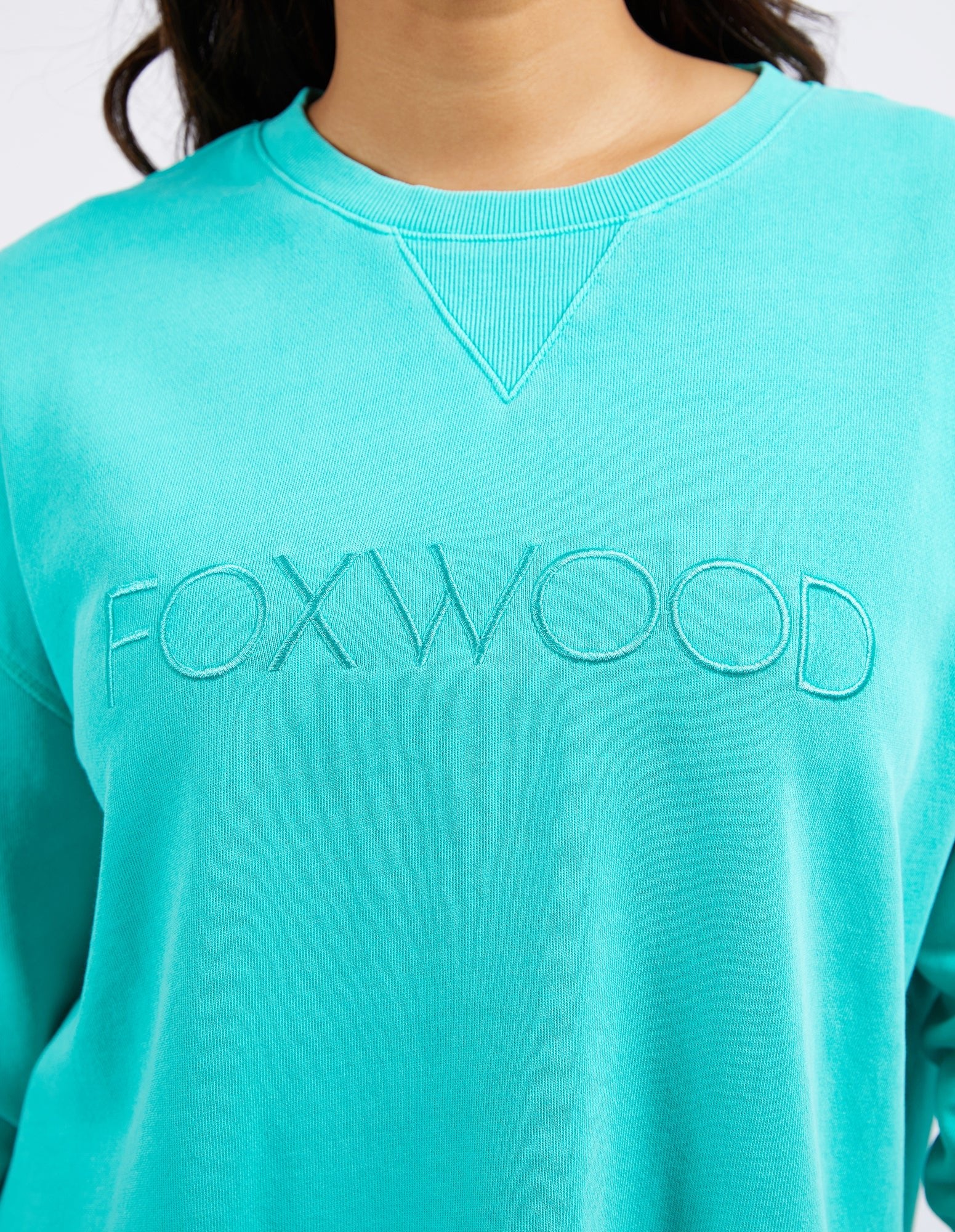 Image of Woman wearing Foxwood Simplified crew in a teal colourway paired with denim jeans on a plain background 
