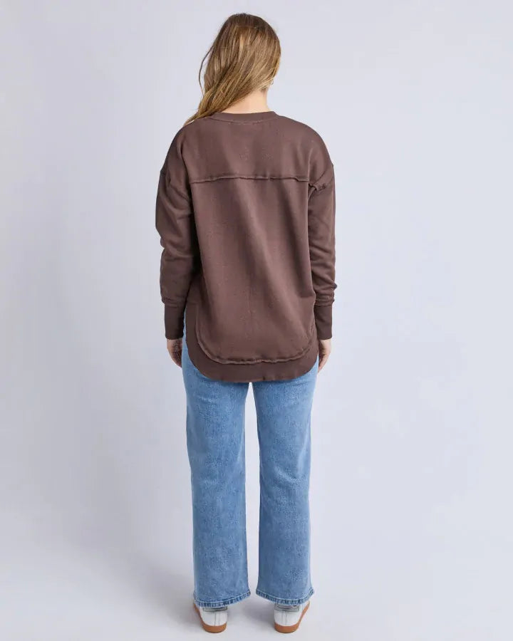 Image of Woman wearing a Foxwood brown sweatshirt and blue jeans on a light gray background