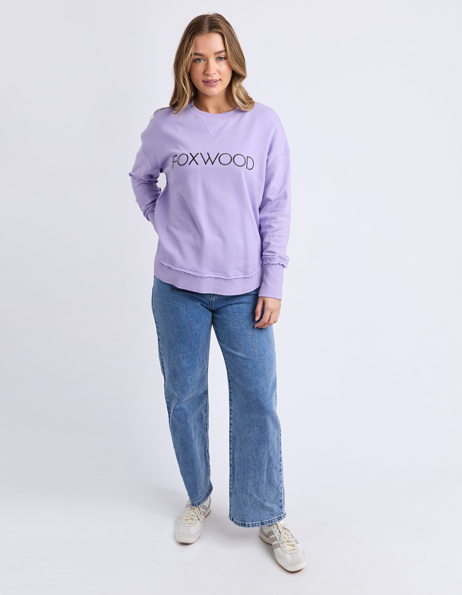 Image of Woman wearing a lavender jumper with 'FOXWOOD' logo and blue jeans on a white background