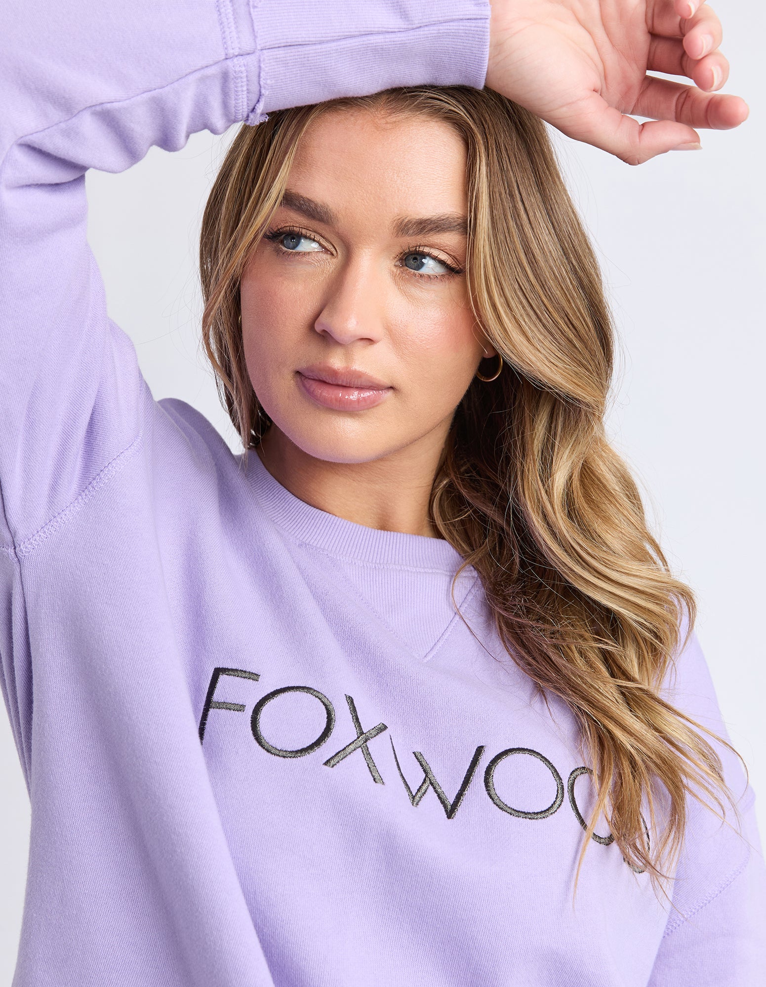 A close up of a Woman wearing a lavender jumper with 'FOXWOOD' text on a plain background