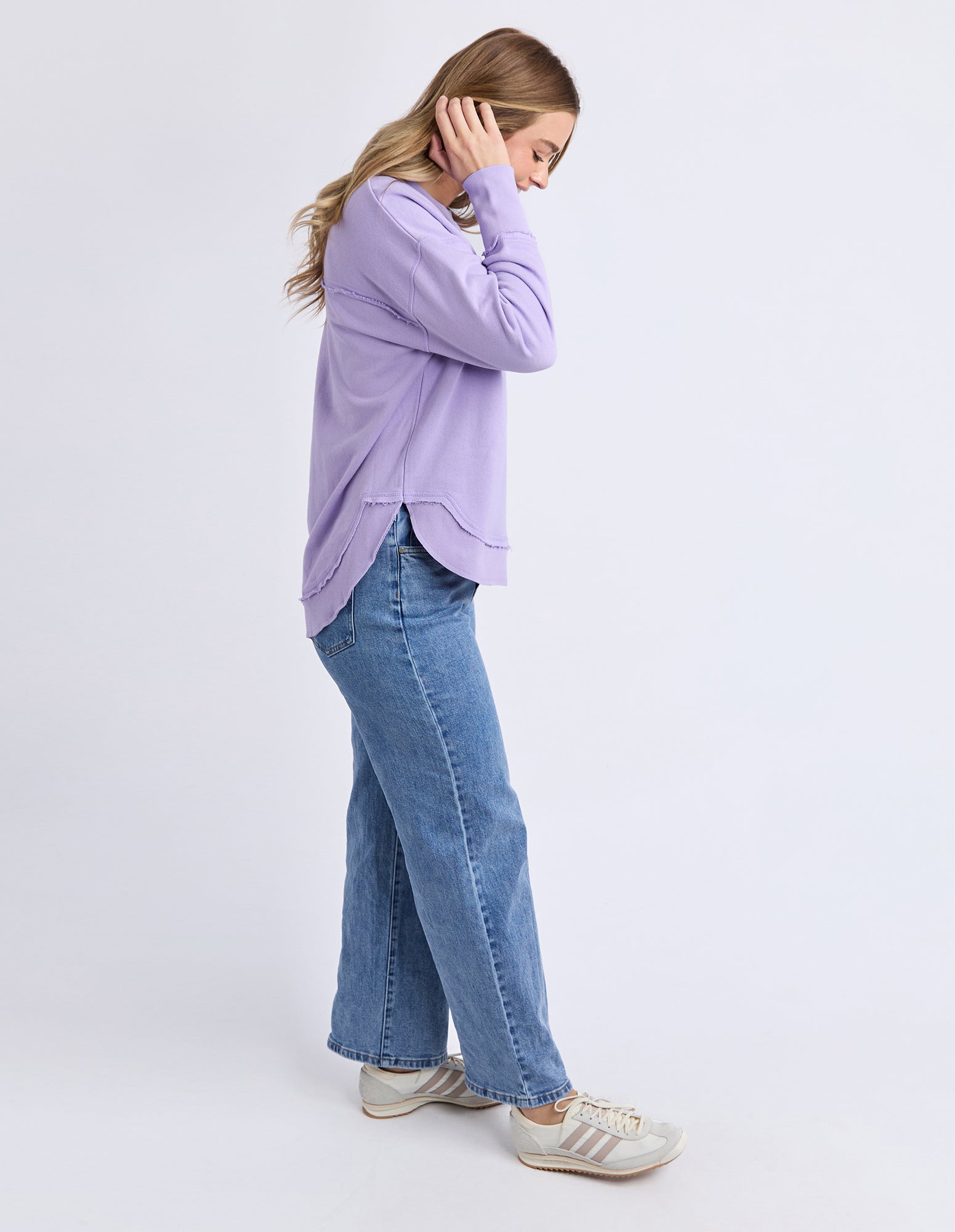 Image of Side view of a Woman wearing a light purple long-sleeve jumper and blue jeans on a white background