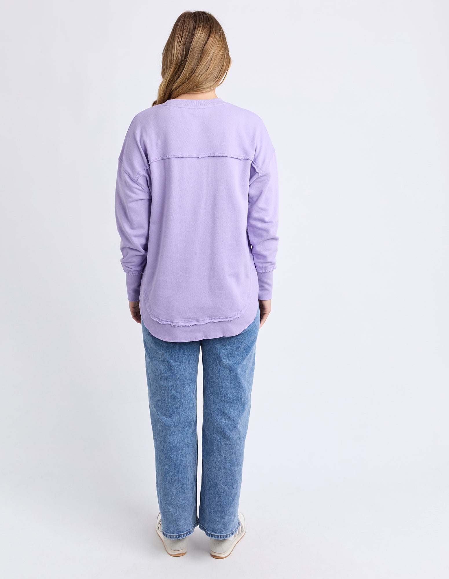 Image of Back view of a woman wearing a light purple/lilac jumper and blue jeans on a white background