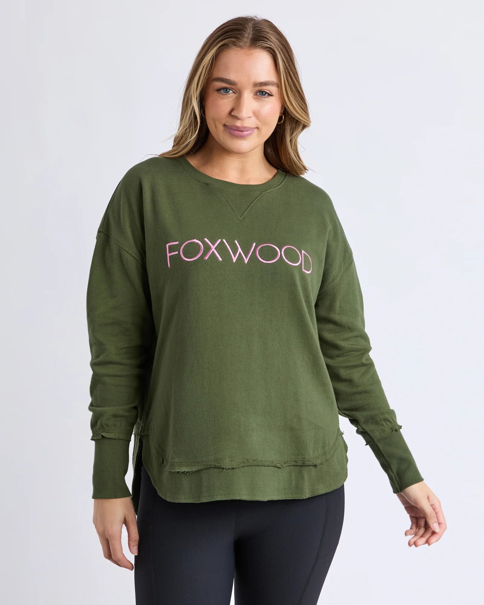 Image of A woman wearing a Foxwood olive green crewneck sweatshirt with 'FOXWOOD' printed in pink across the chest, paired with black leggings, against a light gray studio background.