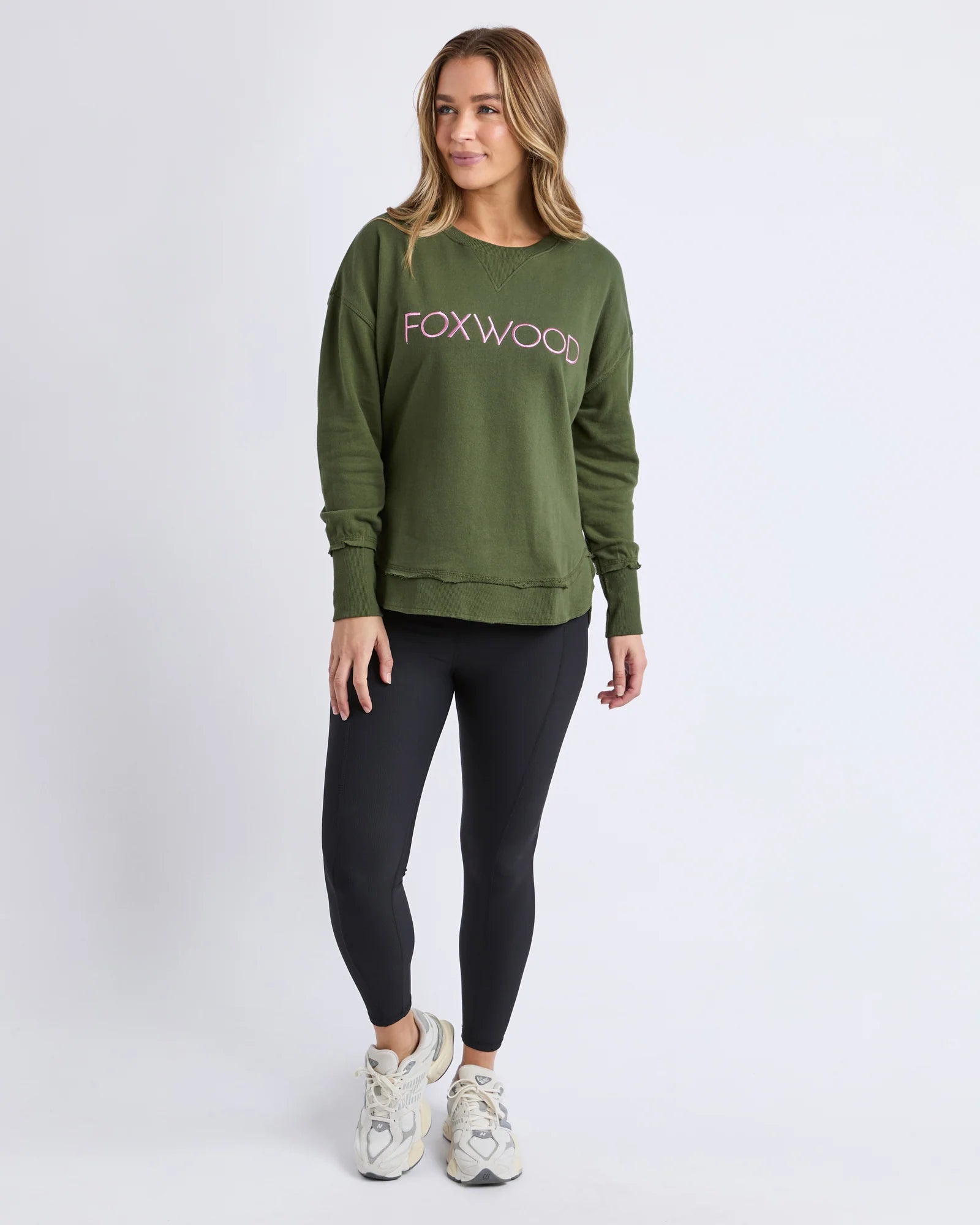 Image of A woman modeling a Foxwood olive green crewneck sweatshirt with 'FOXWOOD' printed across the chest in pink, paired with black leggings and white sneakers, against a light gray studio background.
