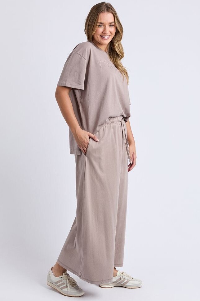 Image of A woman modeling a matching Foxwood taupe two-piece lounge set consisting of a short-sleeve V-neck top and wide-leg drawstring pants, paired with white sneakers, against a light gray studio background.