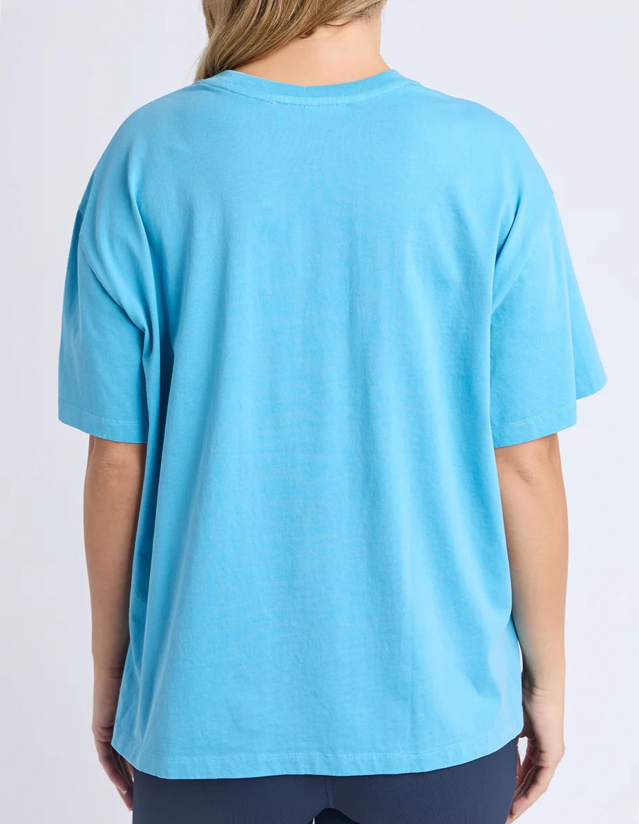 A womanviewed from the back wearing an oversized blue short-sleeve T-shirt against a plain light grey studio background.