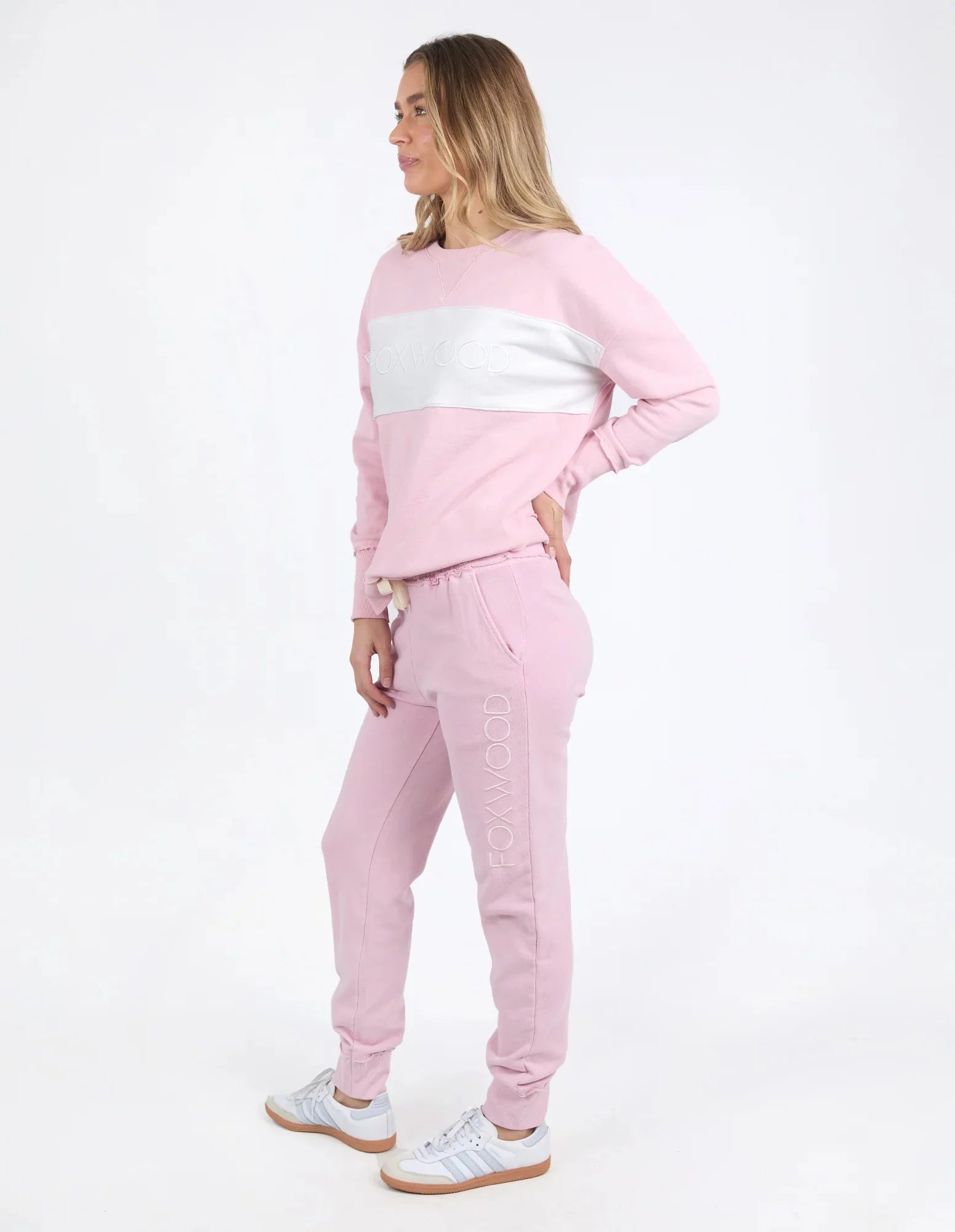 A woman modeling a light pink color-blocked sweatshirt and Foxwood matching jogger pants set, featuring a white chest stripe on the sweatshirt and 'POWEROO' printed vertically on the thigh, against a plain white studio background.