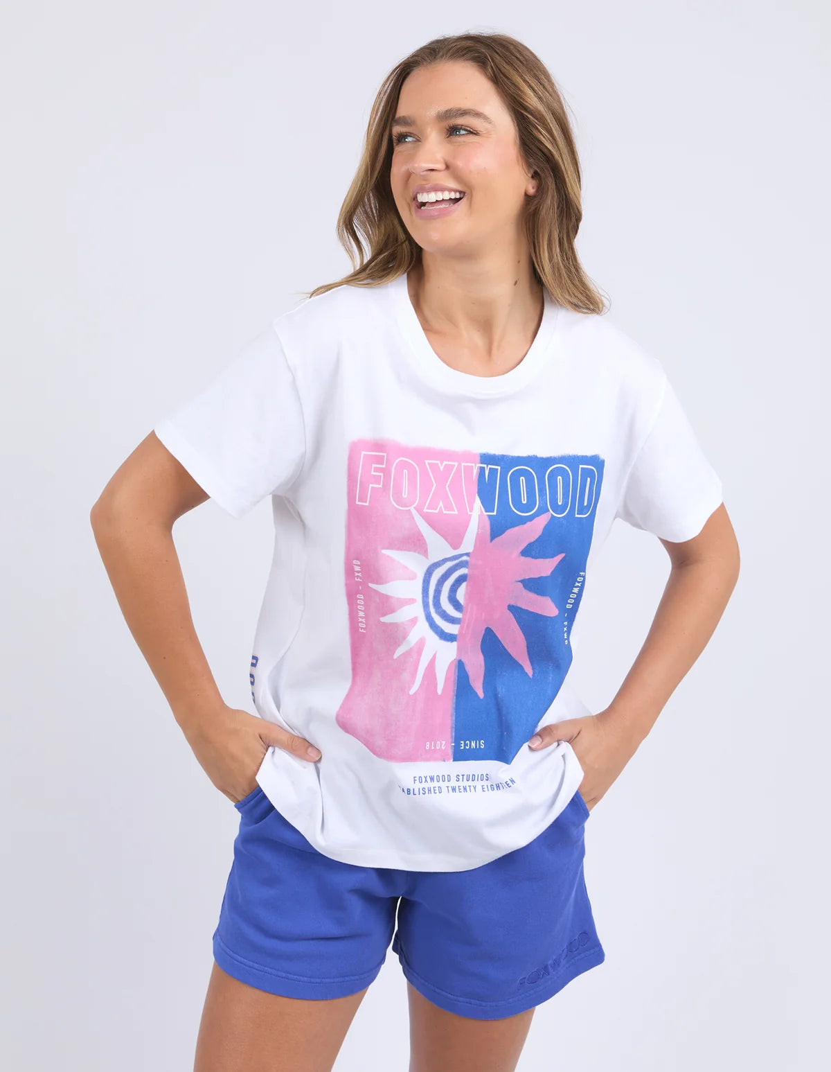 A woman modeling a Foxwood white short-sleeve graphic T-shirt with a pink and blue Foxwood logo print on the front, paired with blue shorts, against a light gray studio background.