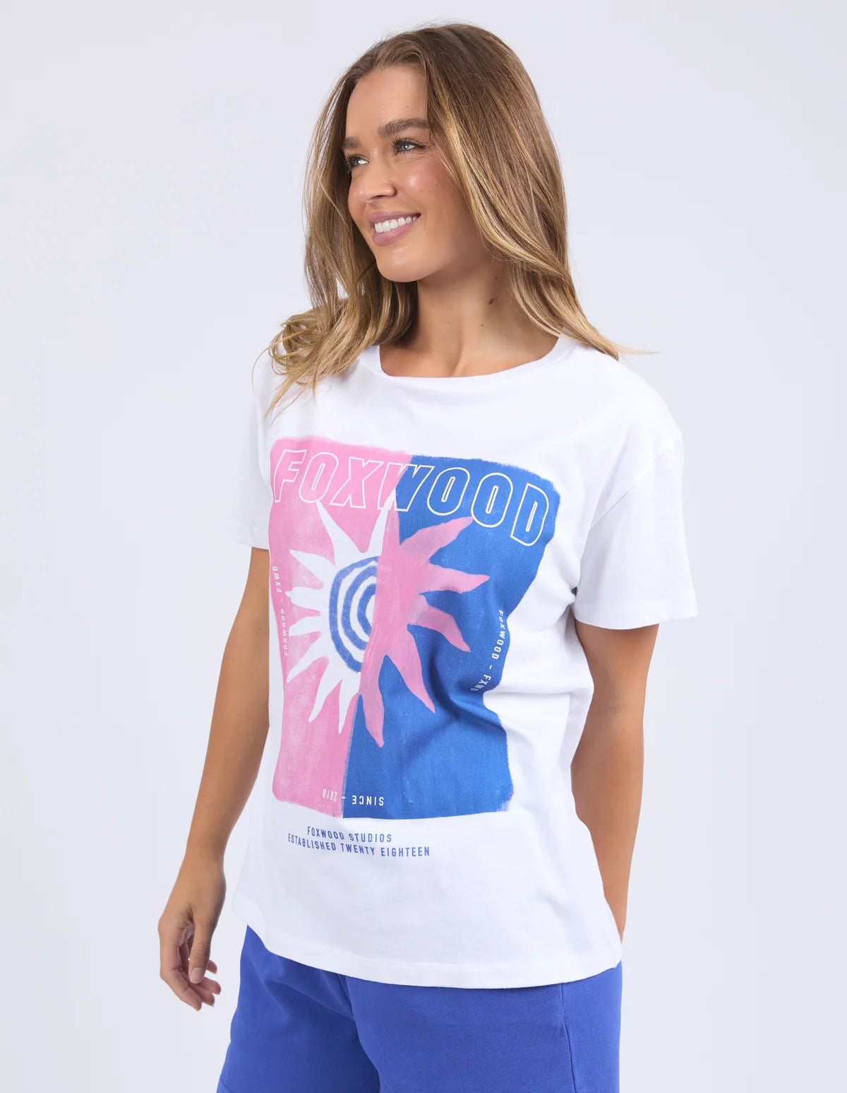 Image of A woman wearing a Foxwood white short-sleeve graphic T-shirt with a pink and blue Foxwood logo print on the front, paired with blue pants, against a light gray studio background.