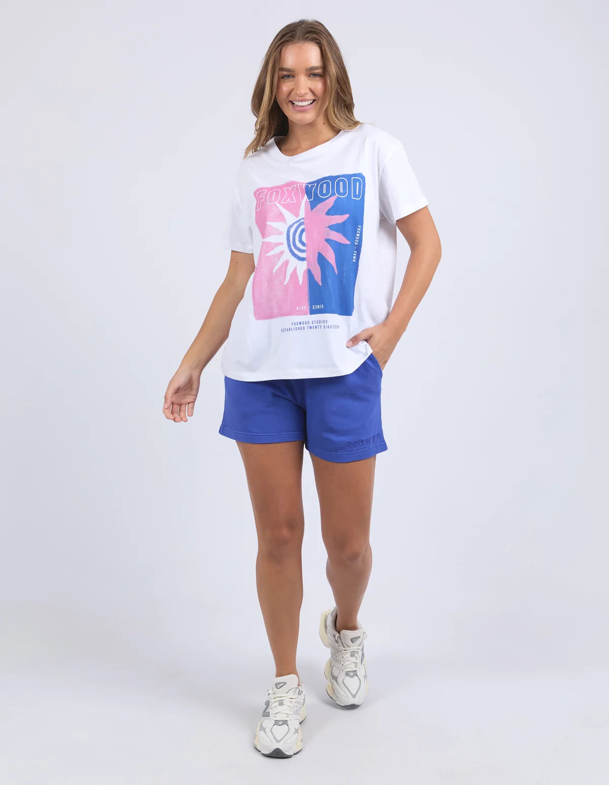 Image of A woman modeling a Foxwood white graphic t-shirt with a pink and blue sun design, paired with blue athletic shorts and white sneakers, against a light gray studio background.