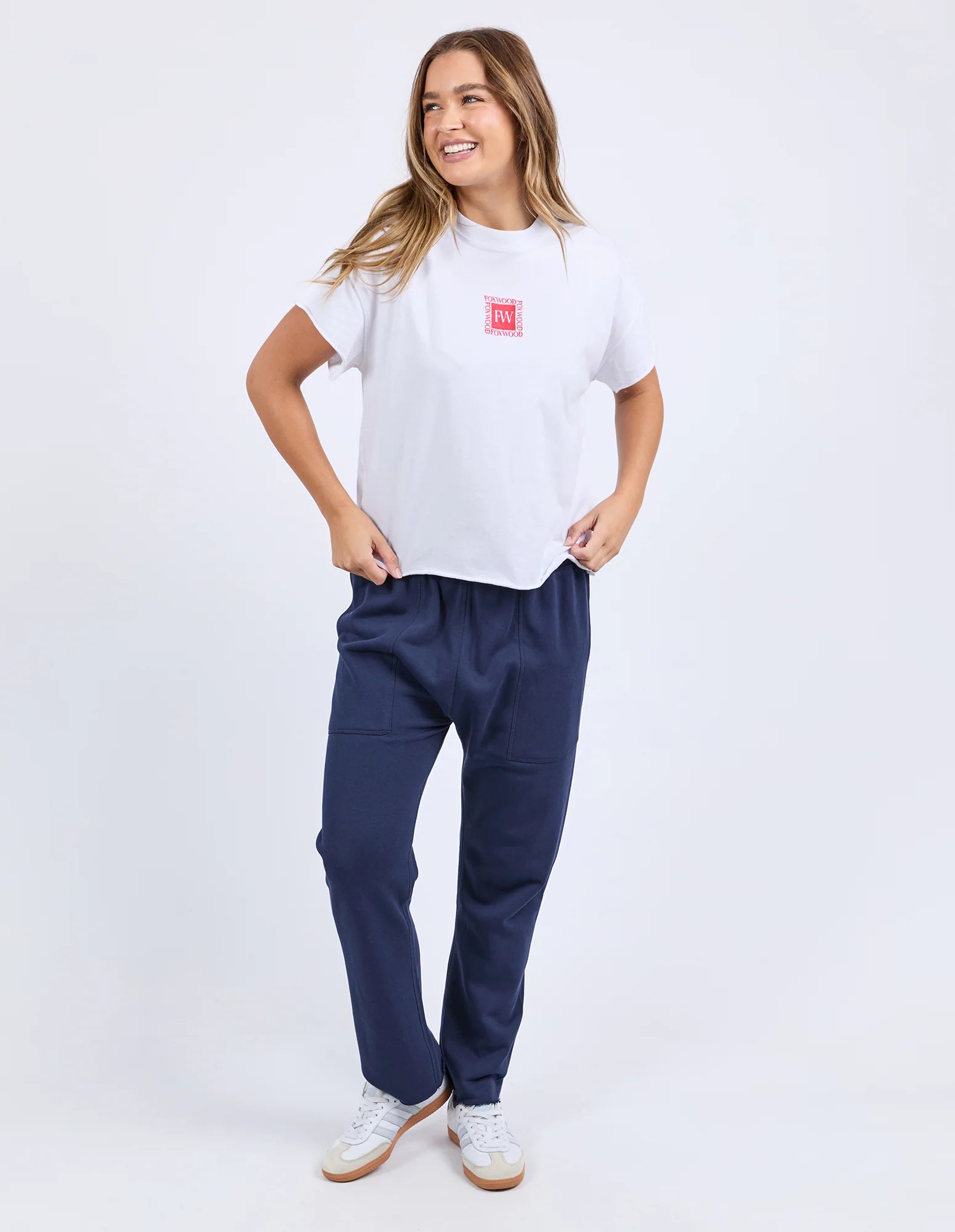 Image of A woman modeling a Foxwood white crewneck t-shirt with a small red square logo on the left chest, paired with navy blue sweatpants and white sneakers, against a plain white studio background.