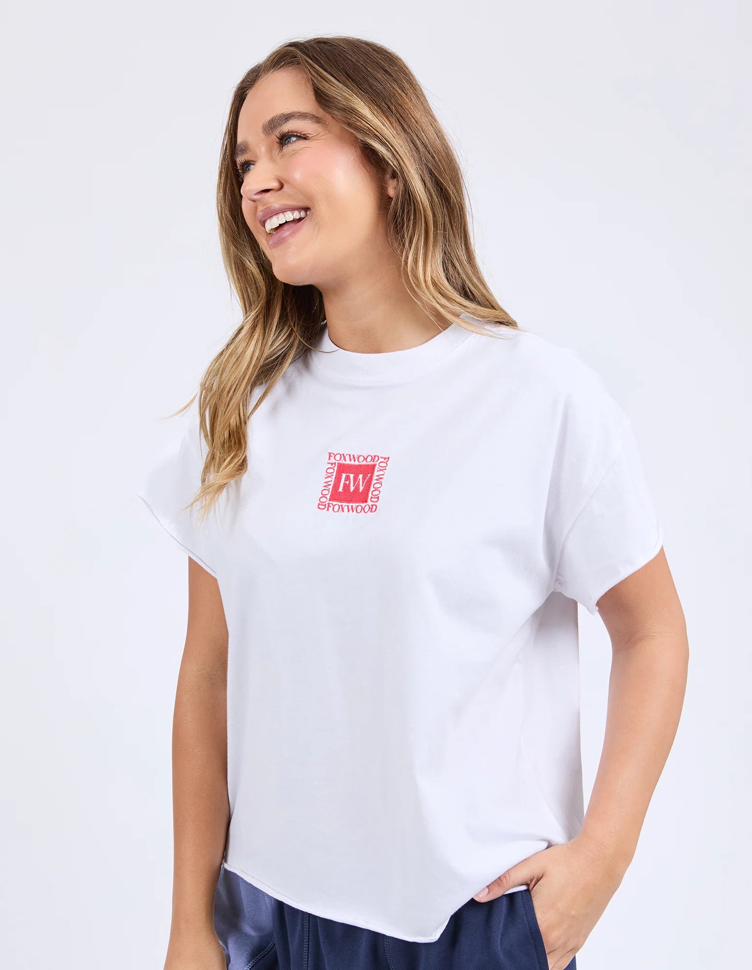 Image of A woman wearing a Foxwood white short-sleeve crewneck t-shirt with a small red and white graphic on the left chest, paired with navy blue pants, set against a plain white studio background.
