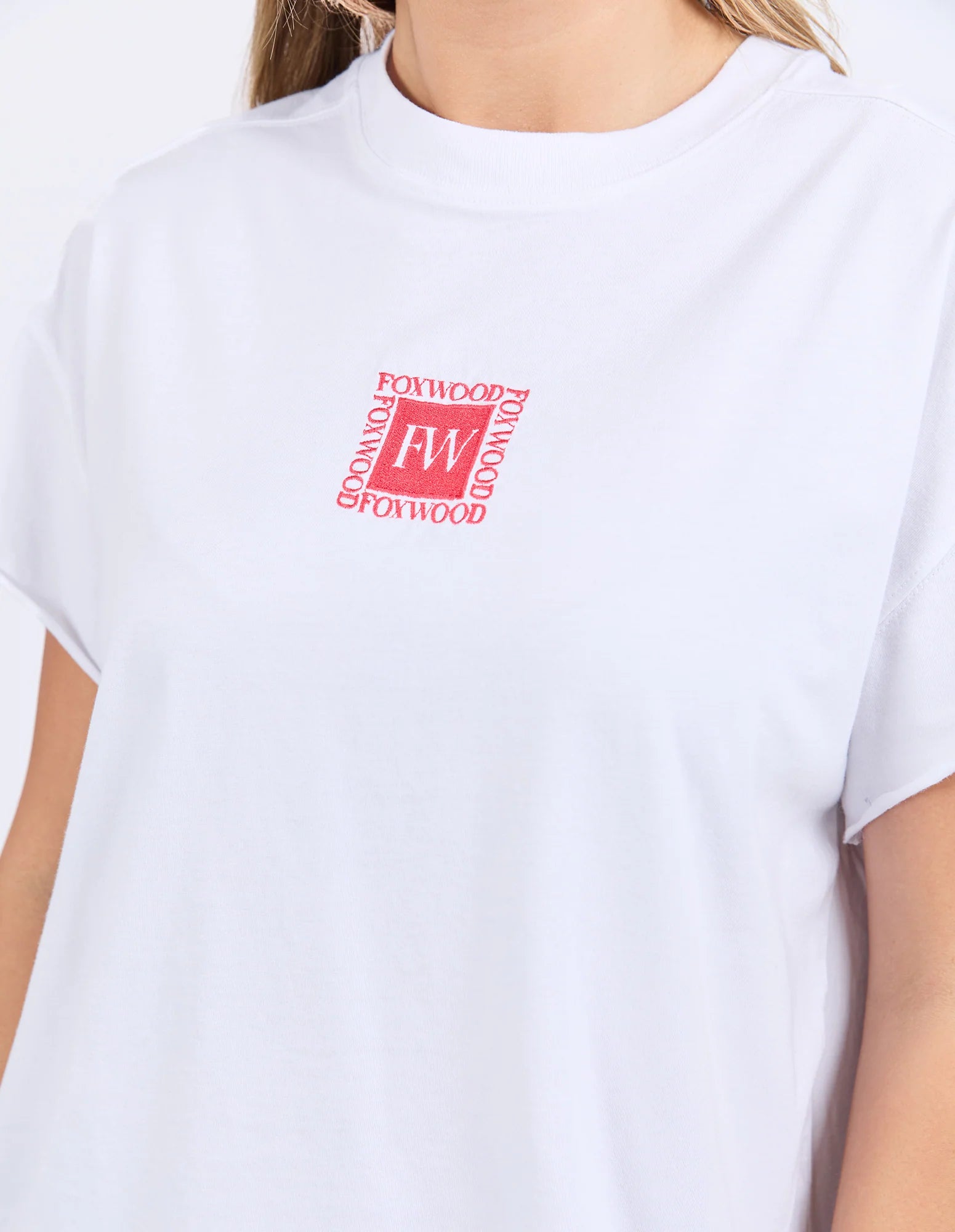A close-up of a woman wearing a Foxwood white short-sleeve crewneck t-shirt with a red square chest graphic that reads 'FORWOOD' around a central 'FW' monogram.
