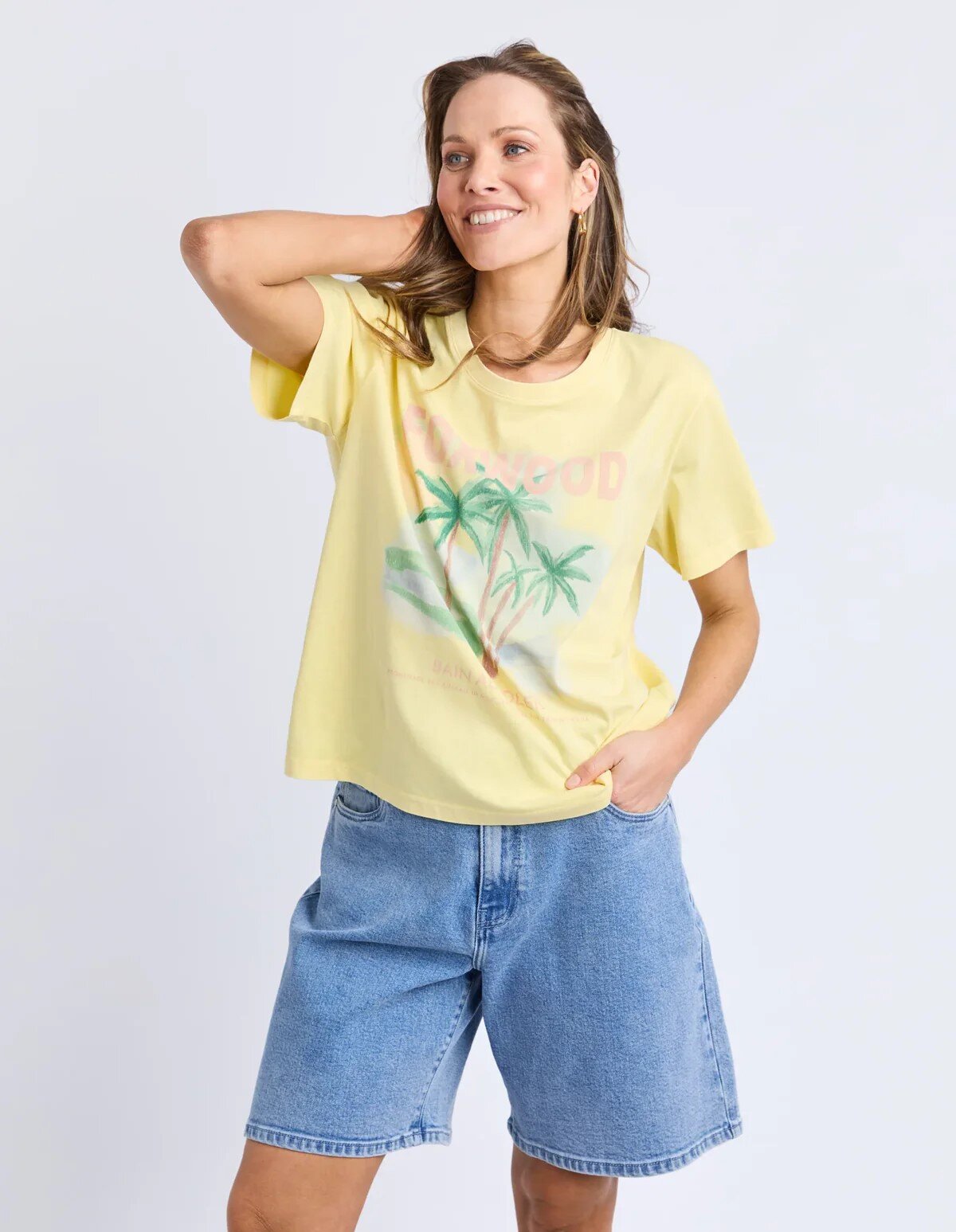 Image of A woman modeling a Foxwood pale yellow graphic t-shirt with a palm tree design, paired with light-wash denim shorts, against a plain light gray studio background.