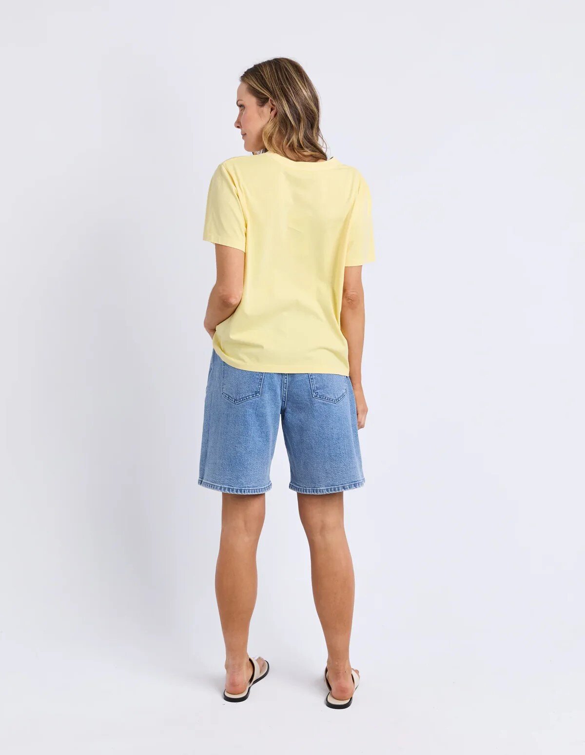 A woman shown from the back wearing a Foxwood pale yellow short-sleeve t-shirt and light blue denim shorts, standing against a plain white studio background.