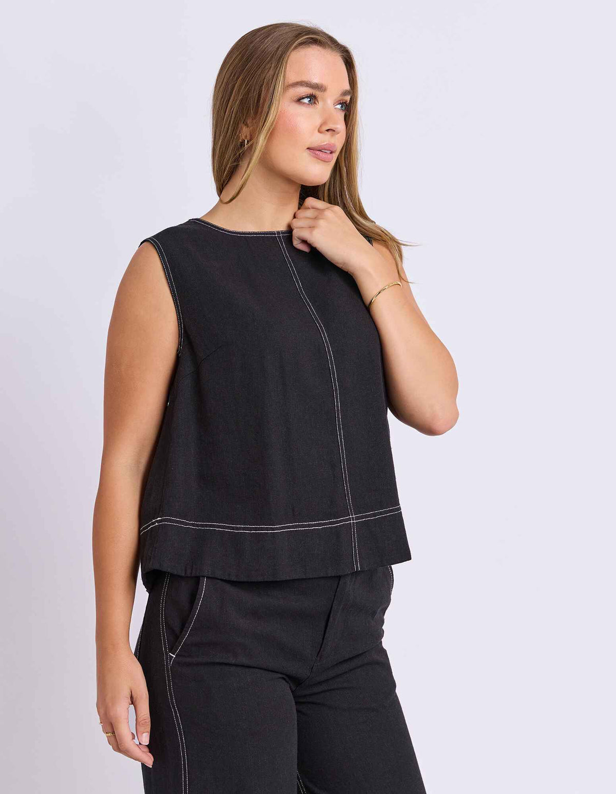 A woman modeling a Foxwood sleeveless black cropped top with contrasting light stitching, paired with black pants, against a light gray studio background.