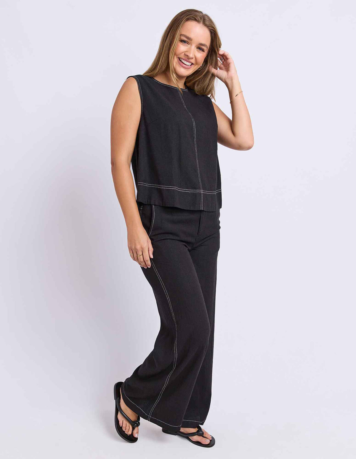 A woman modeling a Foxwood black sleeveless top and matching wide-leg pants with contrasting light stitching, paired with black sandals, against a light gray studio background.