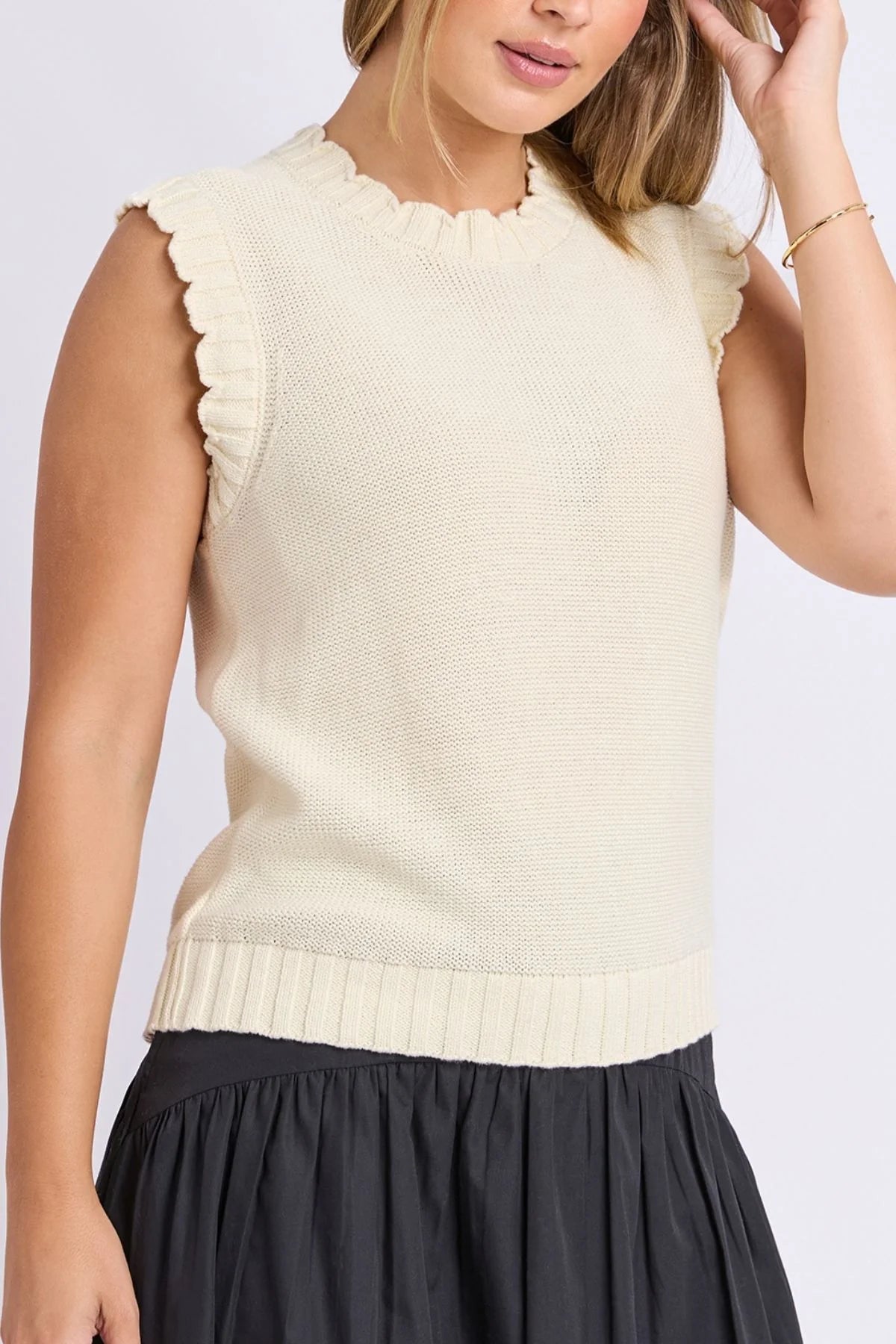 Image of A woman wearing a Foxwood cream-colored sleeveless ribbed knit sweater with scalloped edges and a black skirt, shown from the chest down against a light gray studio background.