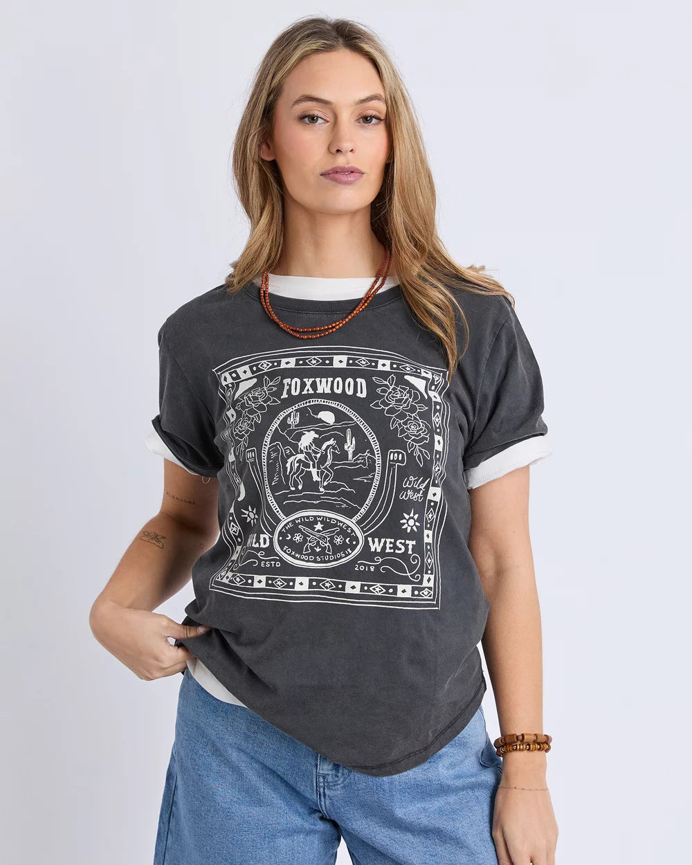 Image of A woman modeling a Foxwood dark gray graphic t-shirt with a white print that reads 'F0XWOOD OLD WESTERN WEST'. The t-shirt has white ruffled trim at the collar and cuffs, paired with blue jeans against a light gray studio background.