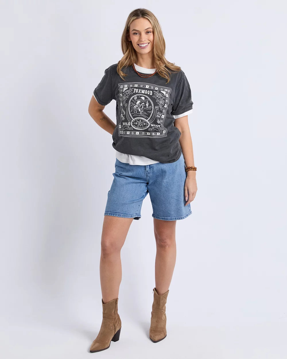 A woman modeling a Foxwood dark gray graphic t-shirt, light blue denim shorts, a white undershirt, and brown ankle boots against a light gray studio background.