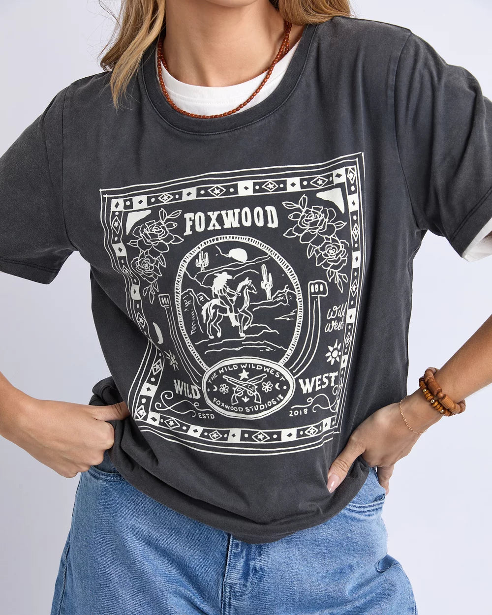 Image of A woman wearing a Foxwood dark charcoal gray graphic T-shirt with a white western-style print reading 'FOXWOOD WEST' and floral/illustrative details, paired with blue jeans against a light gray studio background.