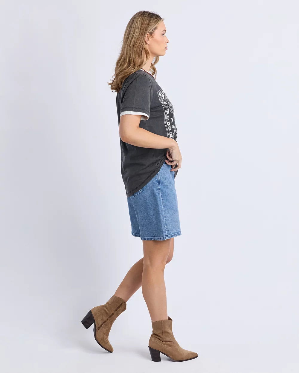 Image of A woman in side profile wearing a dark Foxwood gray graphic t-shirt with white trim, a blue denim skirt, and brown ankle boots, standing against a light gray studio background.