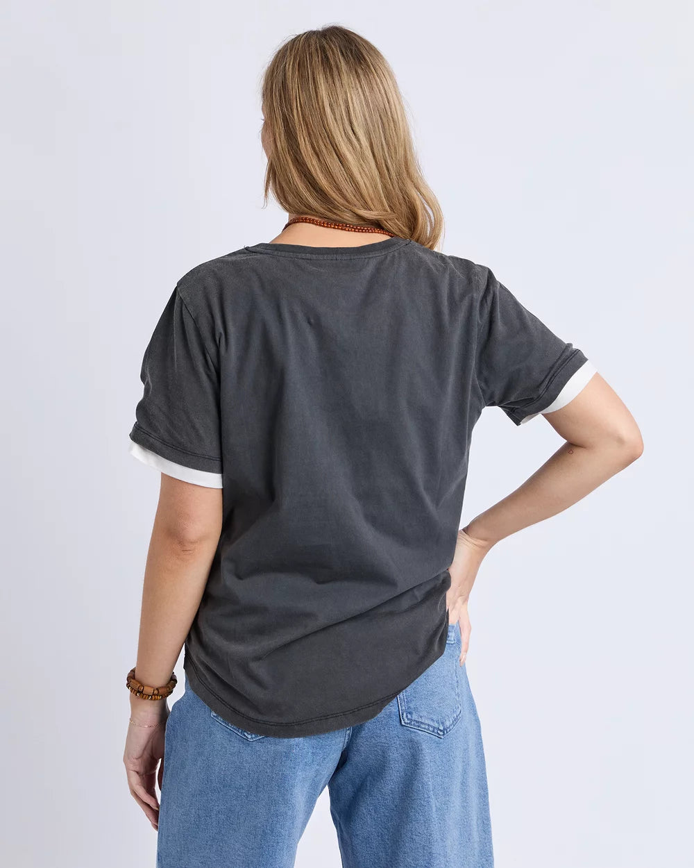 A woman viewed from the back wearing a Foxwood dark charcoal gray ringer t-shirt with white ribbed trim on the sleeves and hem, paired with light blue jeans, against a light gray studio background.