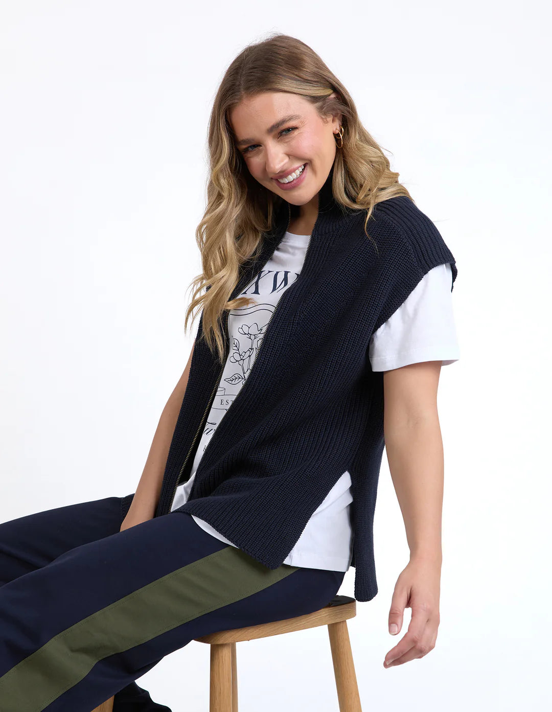 Woman wearing a Foxwood navy blue vest over a white shirt with a graphic design, sitting on a wooden stool against a white background.