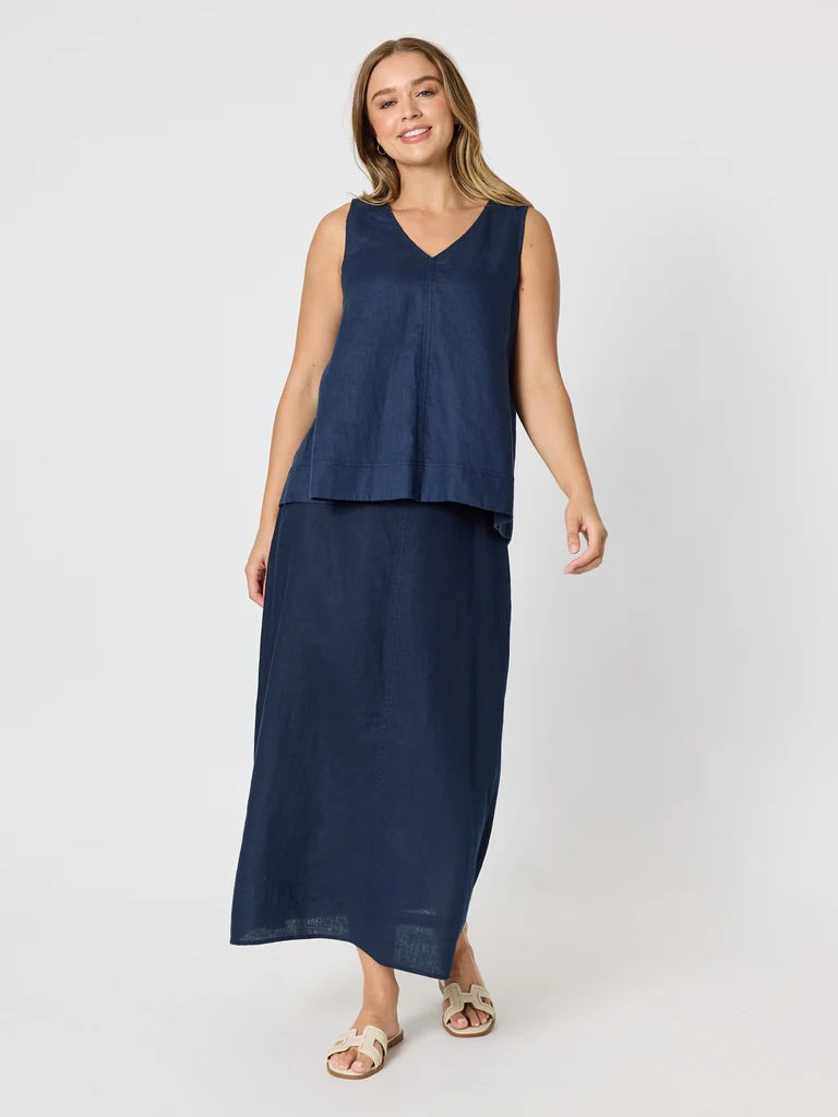 Image of A woman modeling a Gordon Smith sleeveless navy blue two-piece set consisting of a V-neck top and a matching long skirt, paired with beige slide sandals, against a light gray studio background.