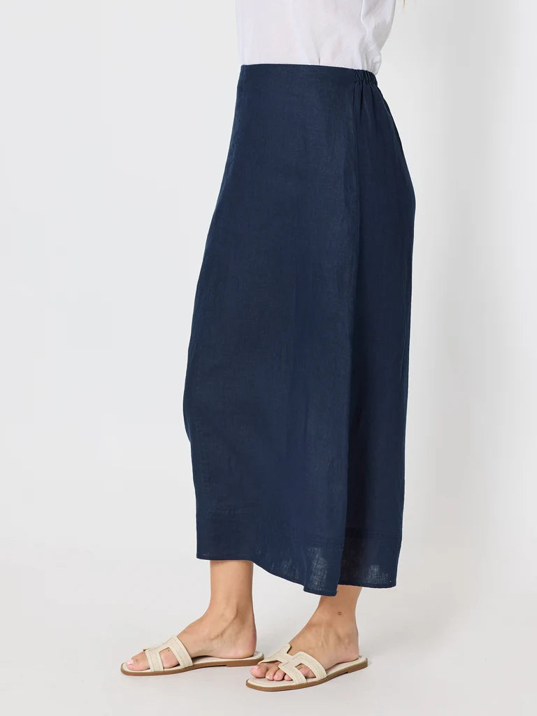 Image of A woman wearing a Gordon Smith navy blue wide-leg cropped skirt or drape-style bottom, paired with a white top and beige slide sandals, shown against a plain light gray studio background.