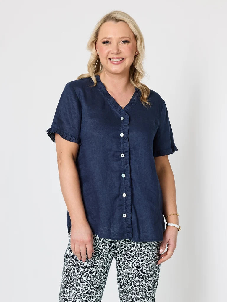 Image of A woman modeling a Gordon Smith navy blue short-sleeve button-up shirt with ruffled trim on the sleeves and collar, paired with patterned pants, against a light gray studio background.