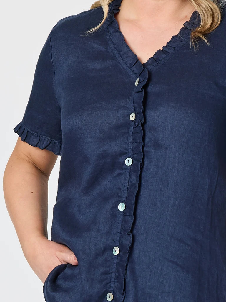 Image of A close-up of a woman wearing a Gordon Smith navy blue short-sleeve button-front top with ruffled trim along the neckline, front placket, and sleeve cuffs, against a light gray background.