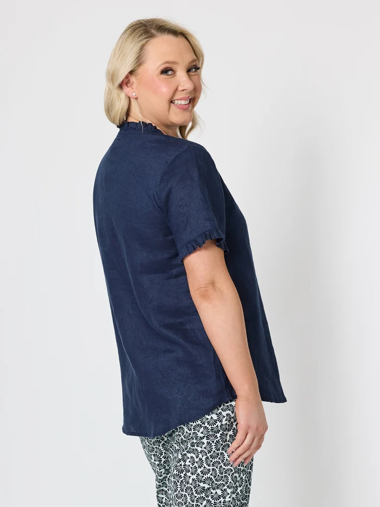 A woman modeling a Gordon Smith navy blue short-sleeve top with a high mock neck and frayed sleeve edges, paired with patterned pants, against a light gray studio background.