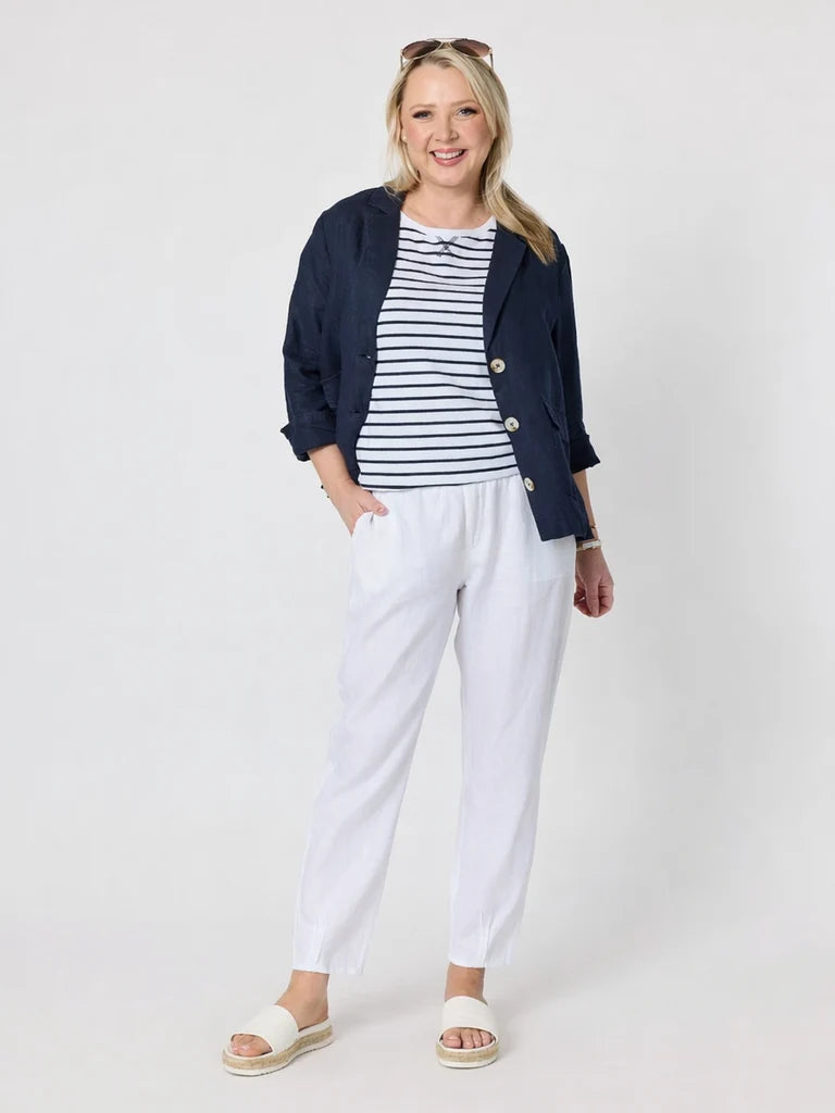 A woman modeling a Gordon Smith casual outfit consisting of a navy blue blazer, a navy and white striped t-shirt, white trousers, and white slide sandals, against a light gray studio background.