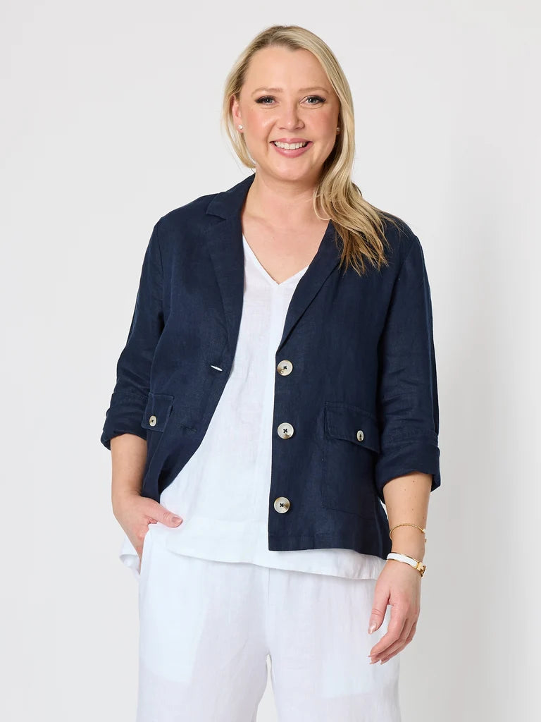 A smiling blonde woman wearing a Gordon Smith navy blue linen-blend blazer with gold-tone buttons over a white V-neck top and white pants, standing against a light gray studio background.