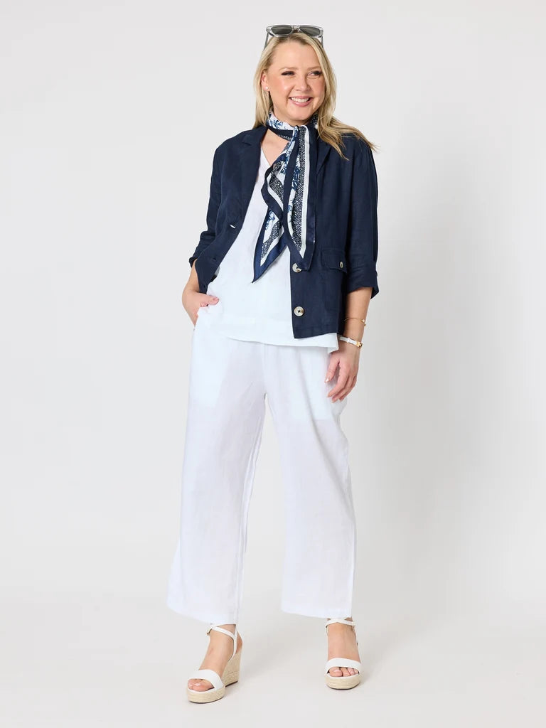 Image of A woman modeling a casual outfit consisting of a Gordon Smith navy blue blazer, white wide-leg pants, a white top, and a navy and white patterned neck scarf, paired with white wedge sandals against a light gray studio background.