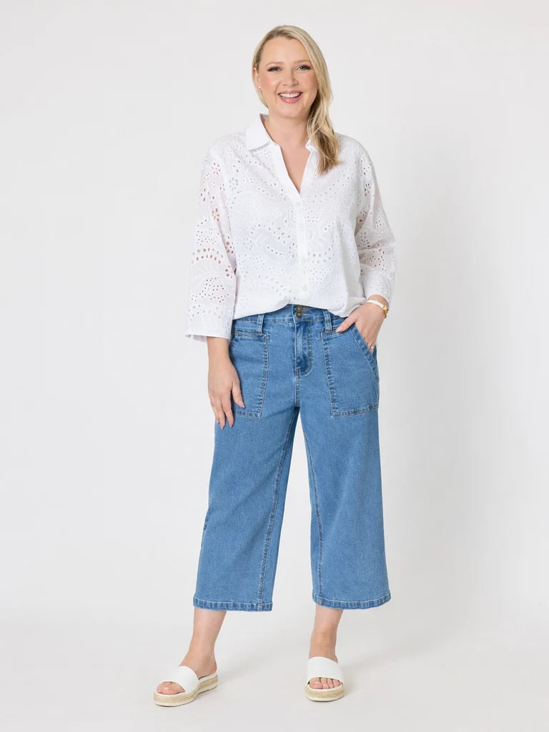 A woman modeling a white eyelet button-up blouse and Gordon Smith blue cropped denim jeans against a plain light gray studio background.