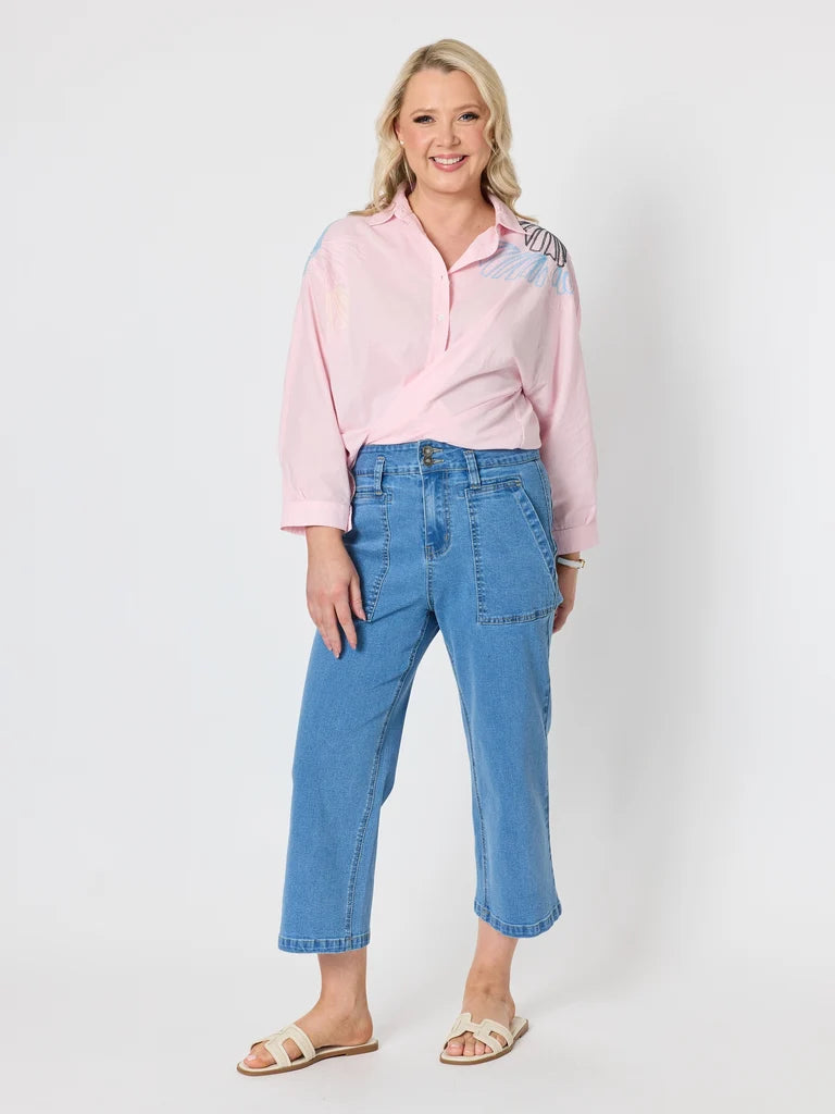 Image of A woman modeling a light pink button-up shirt with a subtle blue and purple gradient on the shoulders, paired with Gordon Smith light-wash blue cropped jeans and beige slide sandals, against a plain light gray studio background.